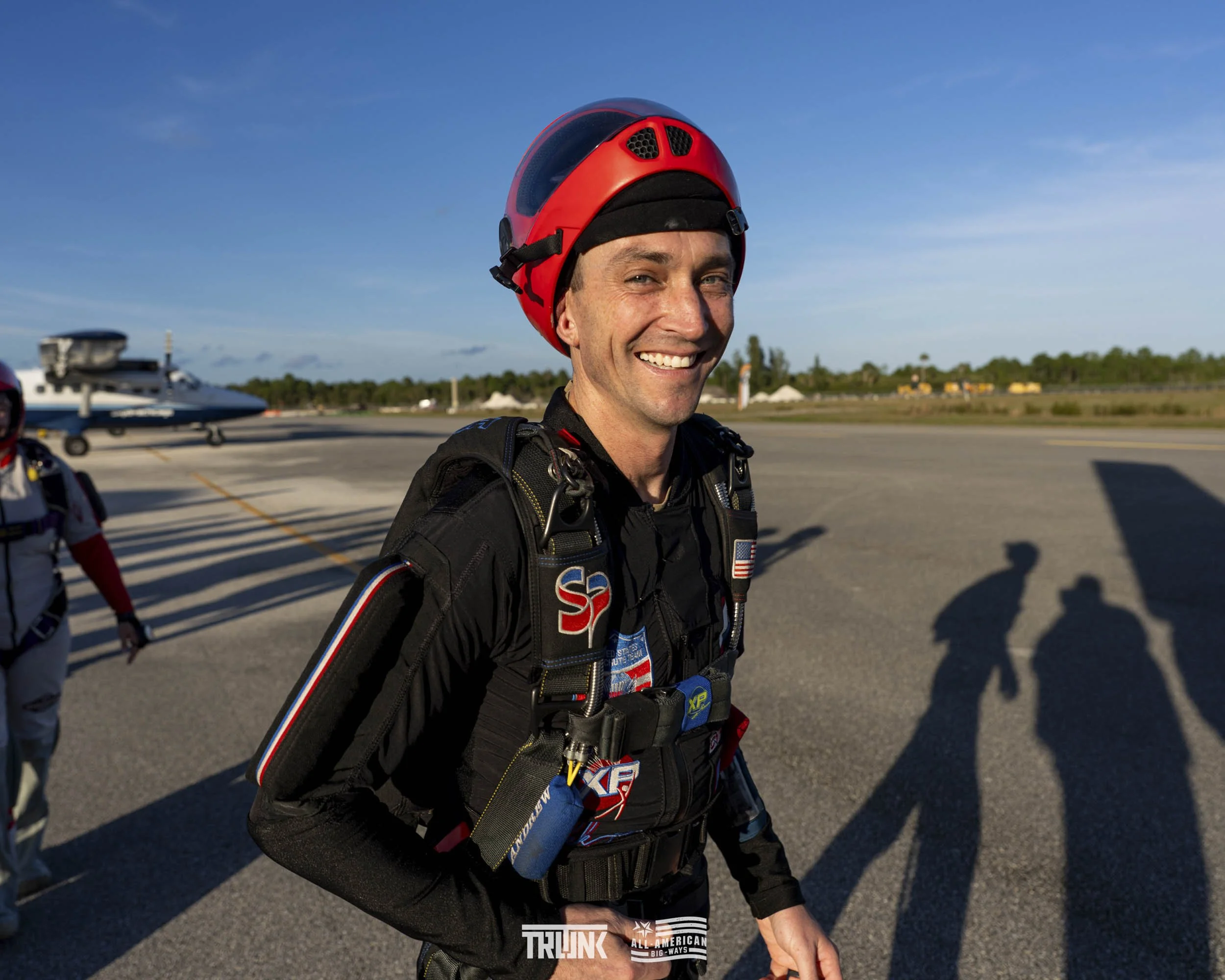 Smiling man in skydiving gear and red helmet on tarmac with shadow, airplane in background, clear sky.