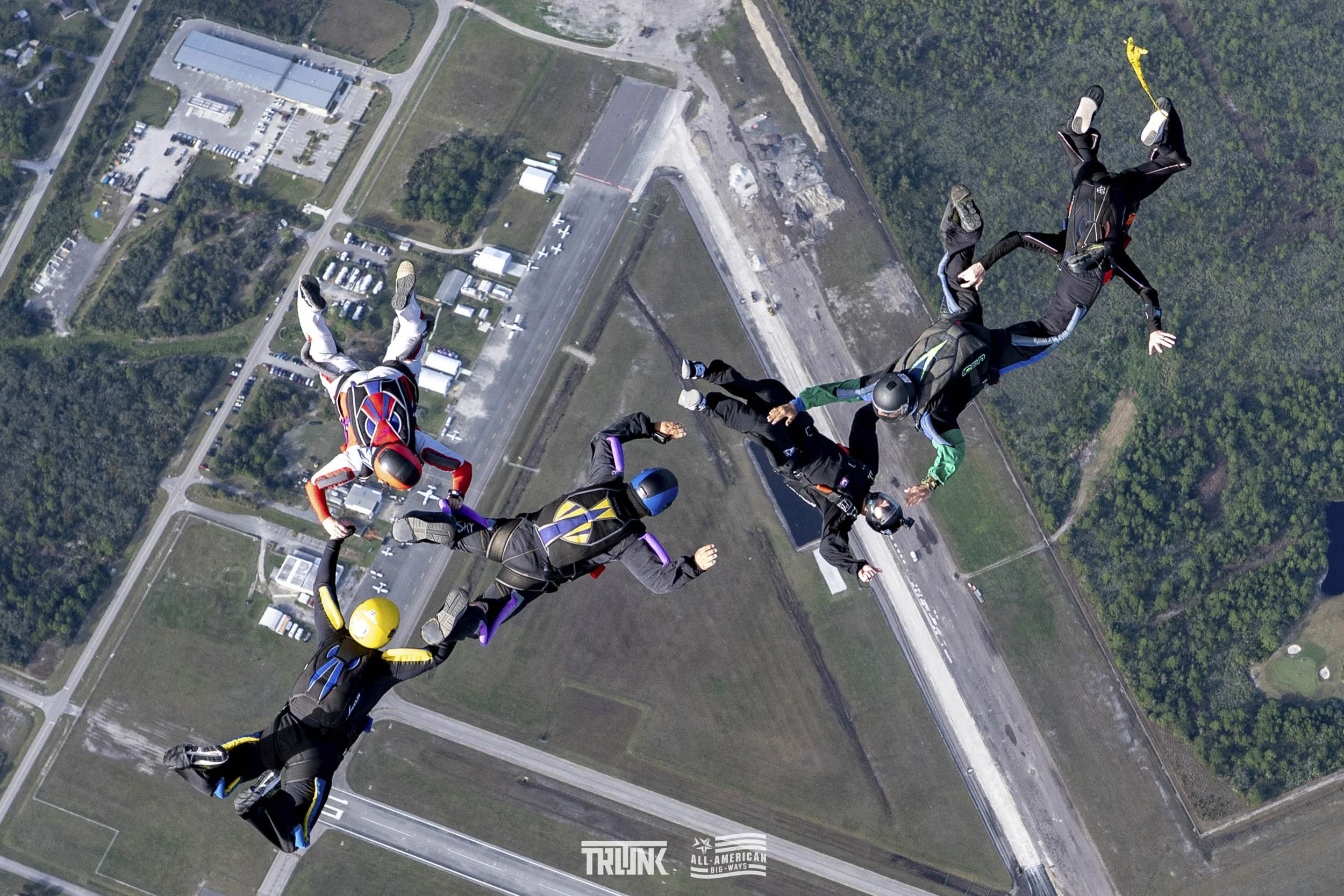 Skydivers in free fall above a landscape with roads, buildings, and trees, holding hands in a formation during a tandem jump.
