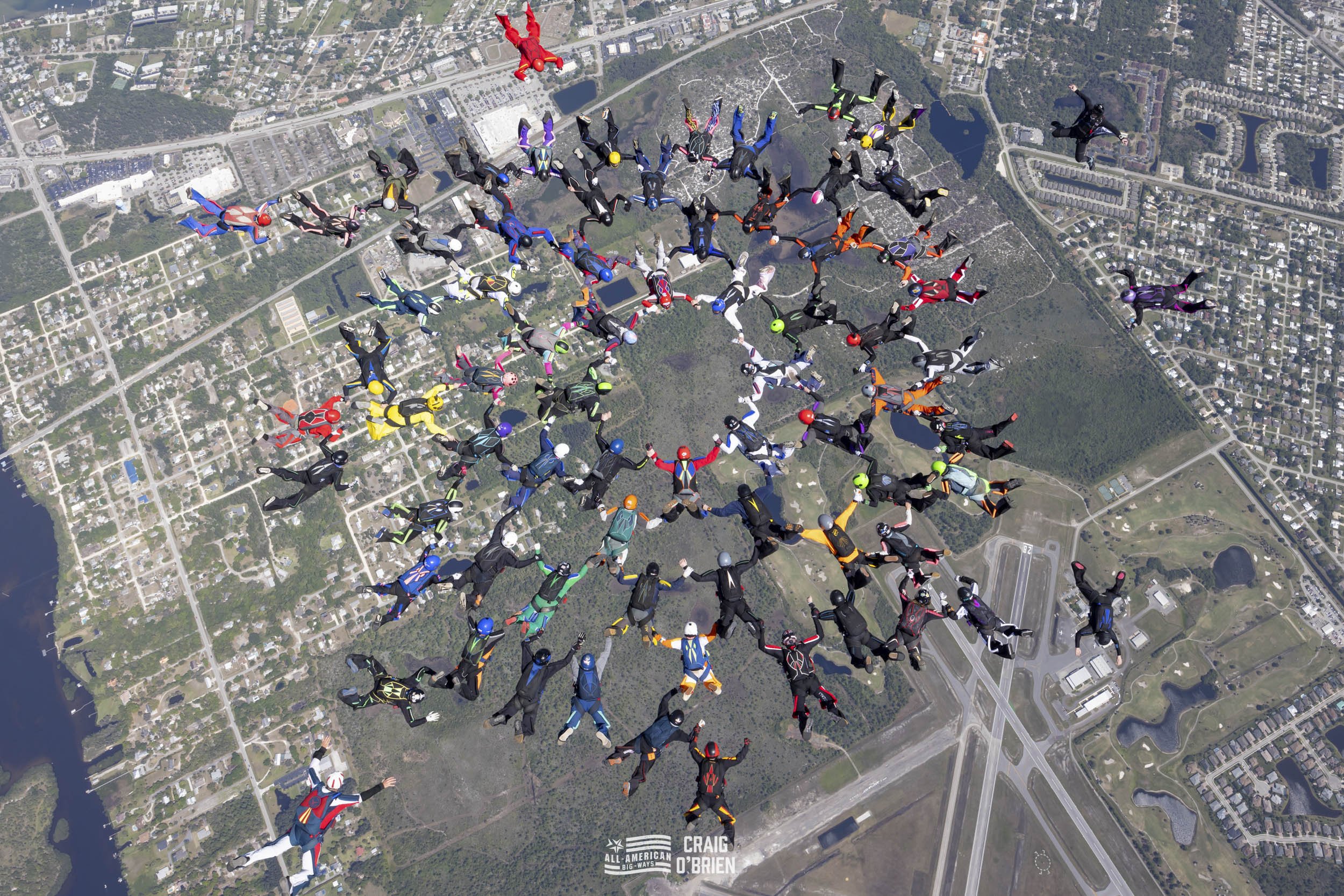A large group of skydivers in colorful jumpsuits and helmets, forming a circle and holding hands while free-falling above a city landscape with roads, lakes, and buildings.