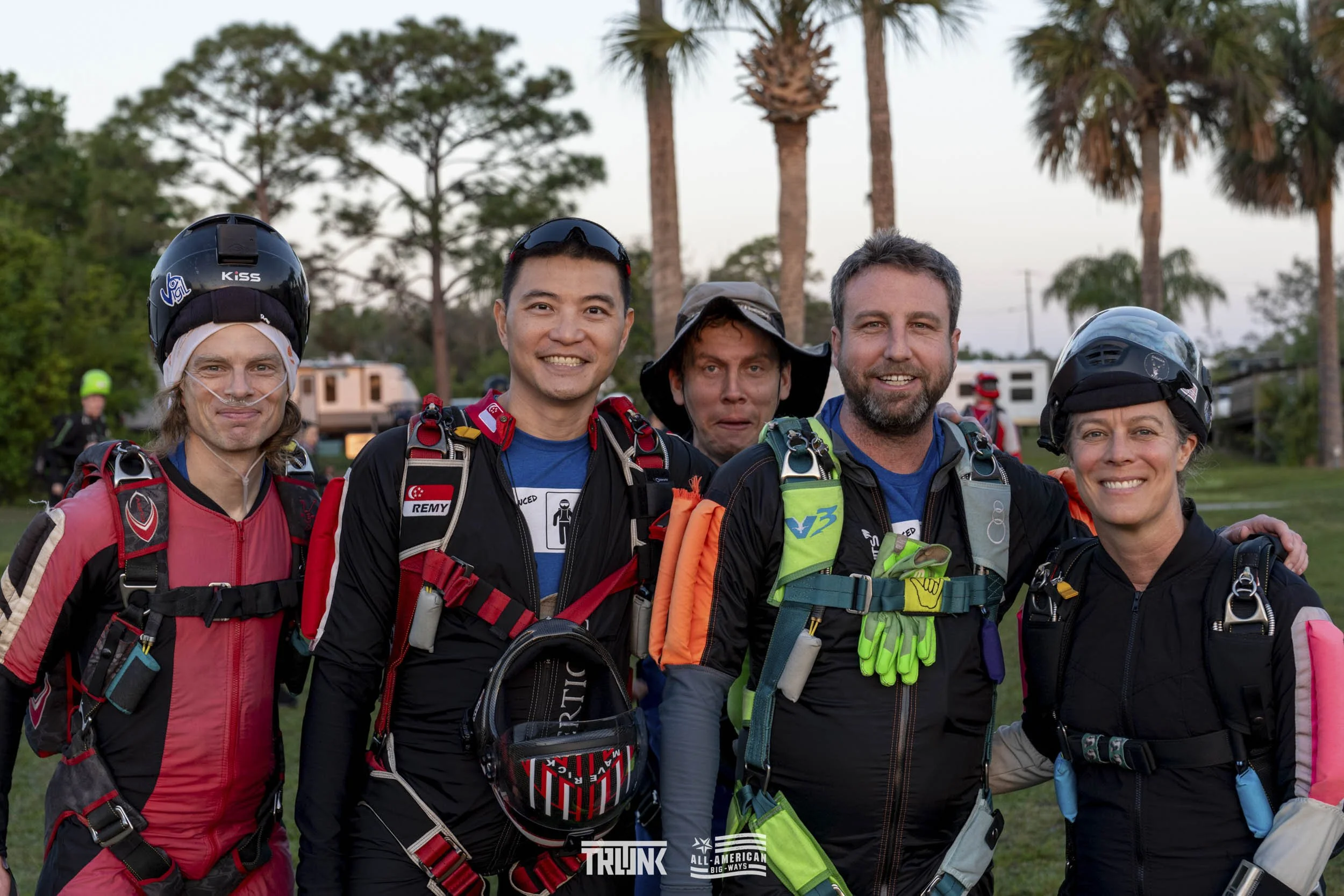 Group of five skydivers in jumpsuits and helmets standing outdoors with palm trees in the background.