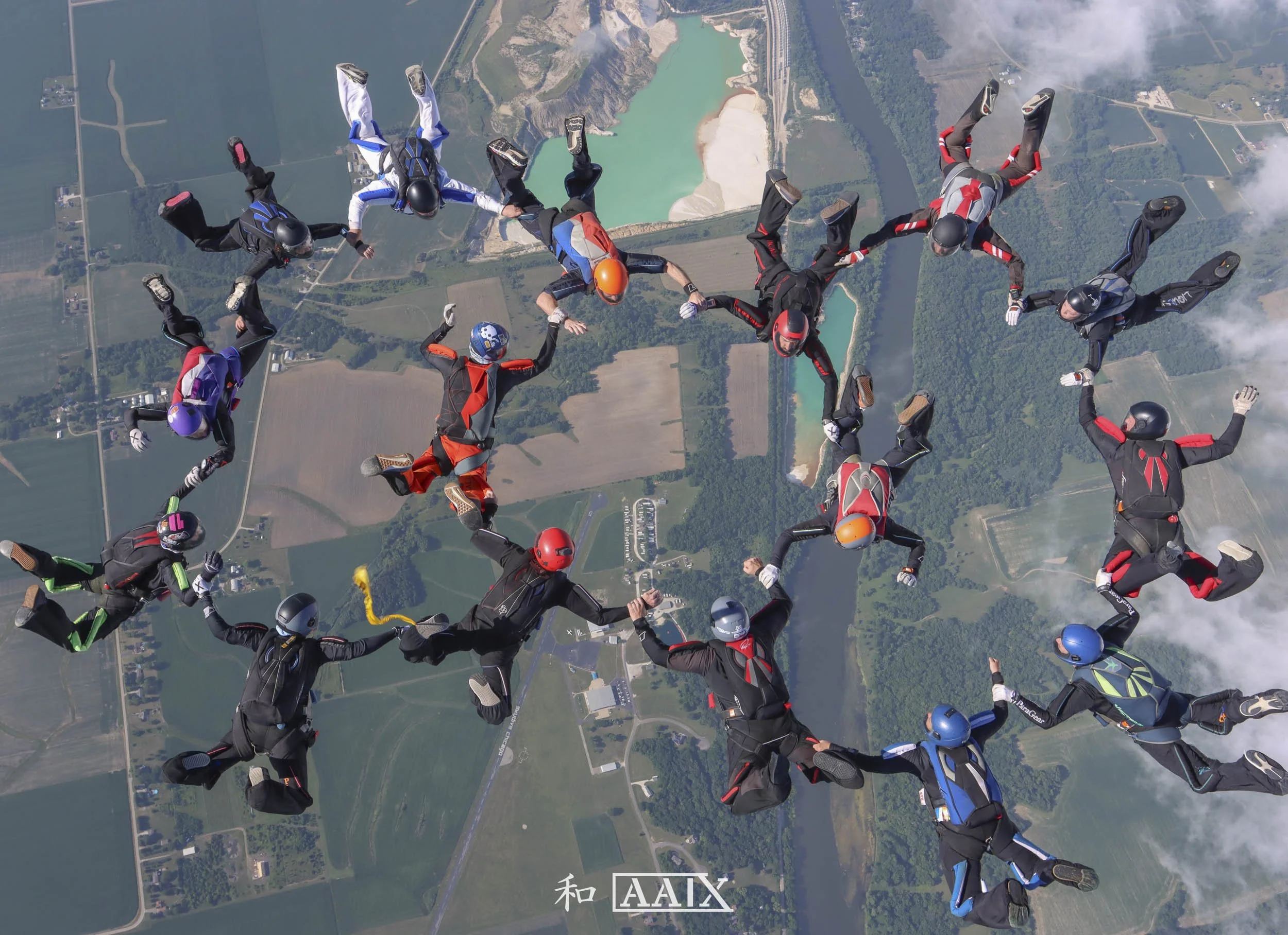 A group of skydivers holding hands in a circle during freefall over a landscape with fields and water bodies.