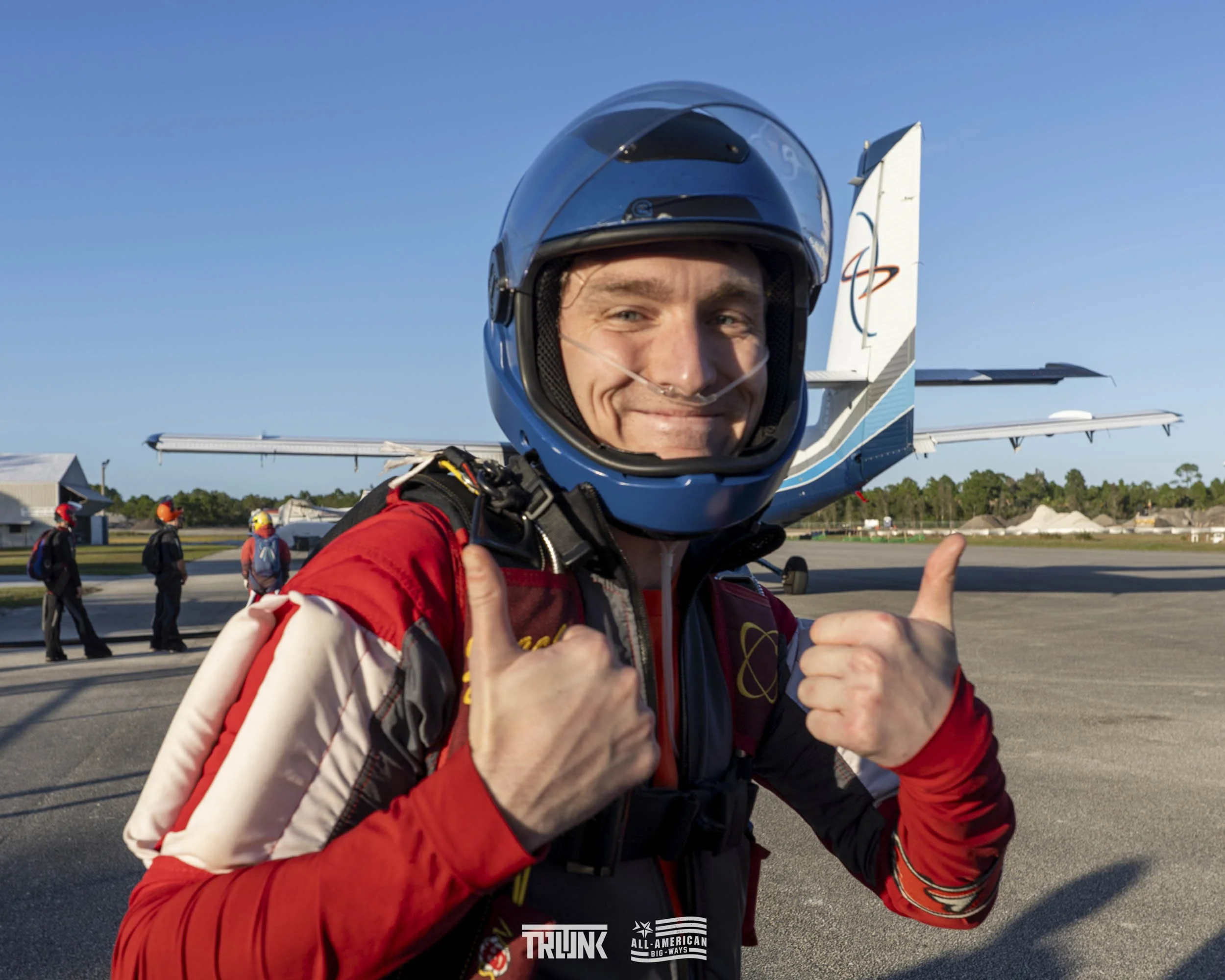 A smiling man in a pilot suit and helmet giving a thumbs-up on an airport tarmac with a small airplane in the background.