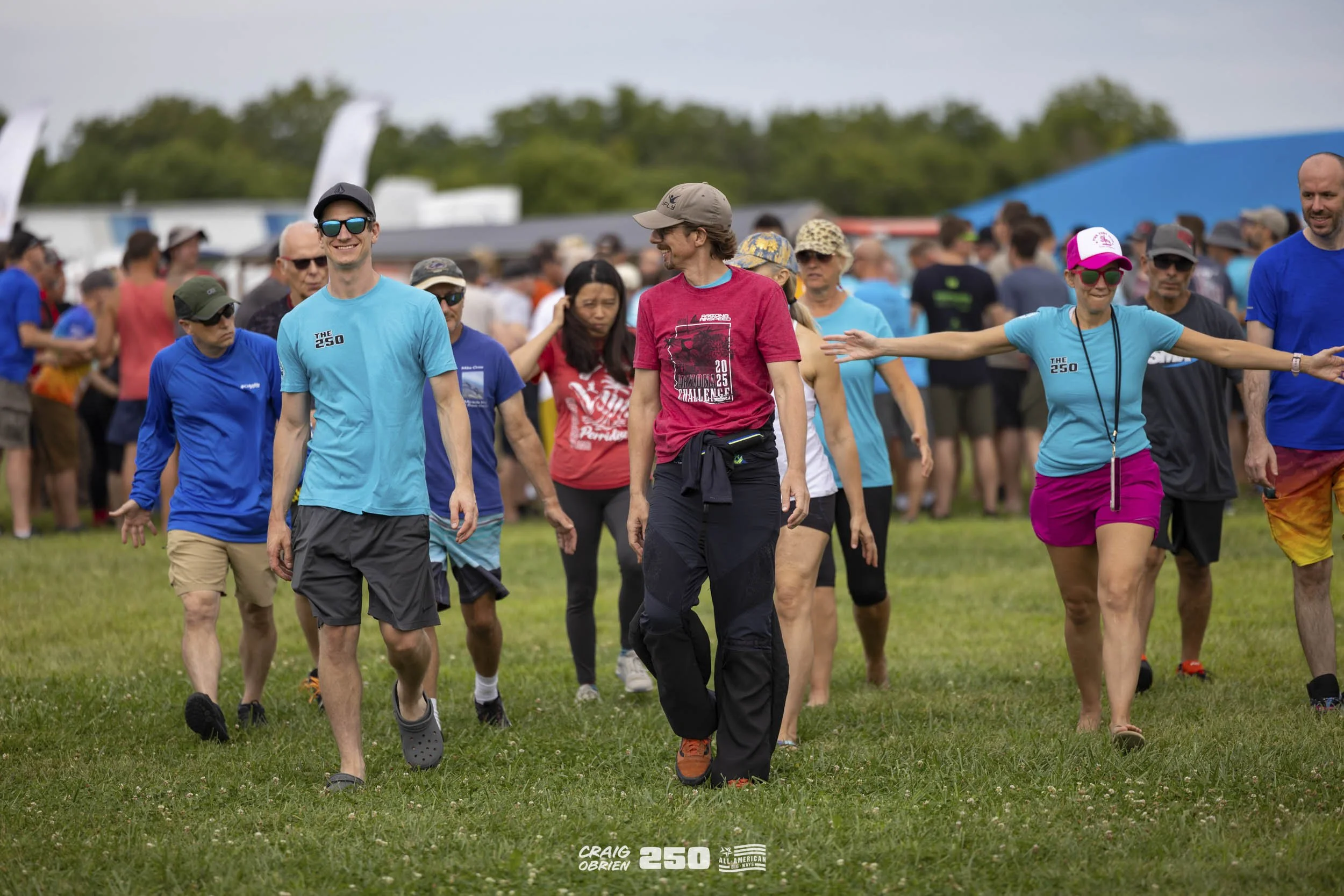 Group of people walking on grass at outdoor event, with some wearing colorful athletic clothing, in front of tents and trees.
