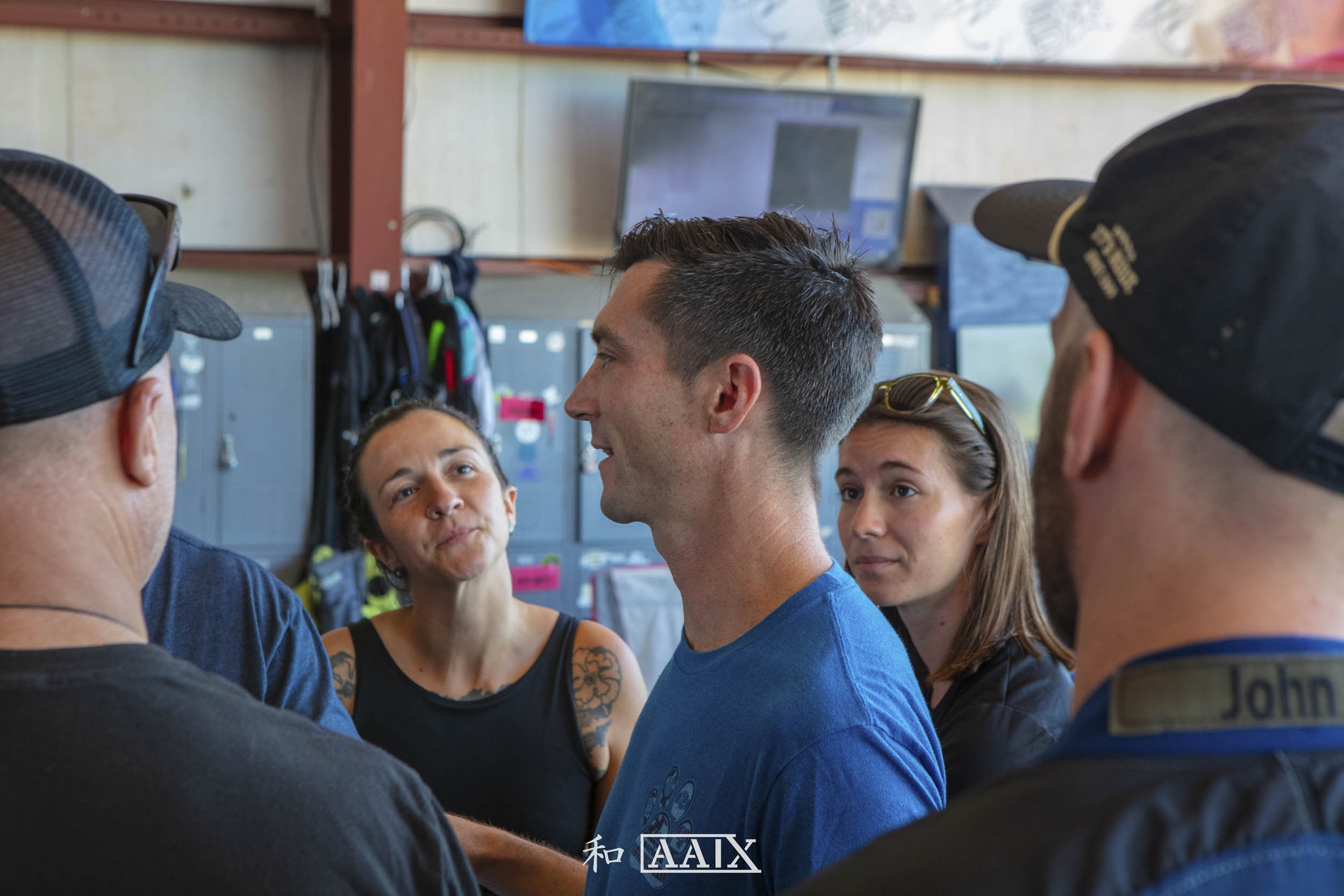 A group of five people in a locker room having a conversation. One man is in the center, facing a woman, with others around him. Lockers and a computer monitor are in the background.