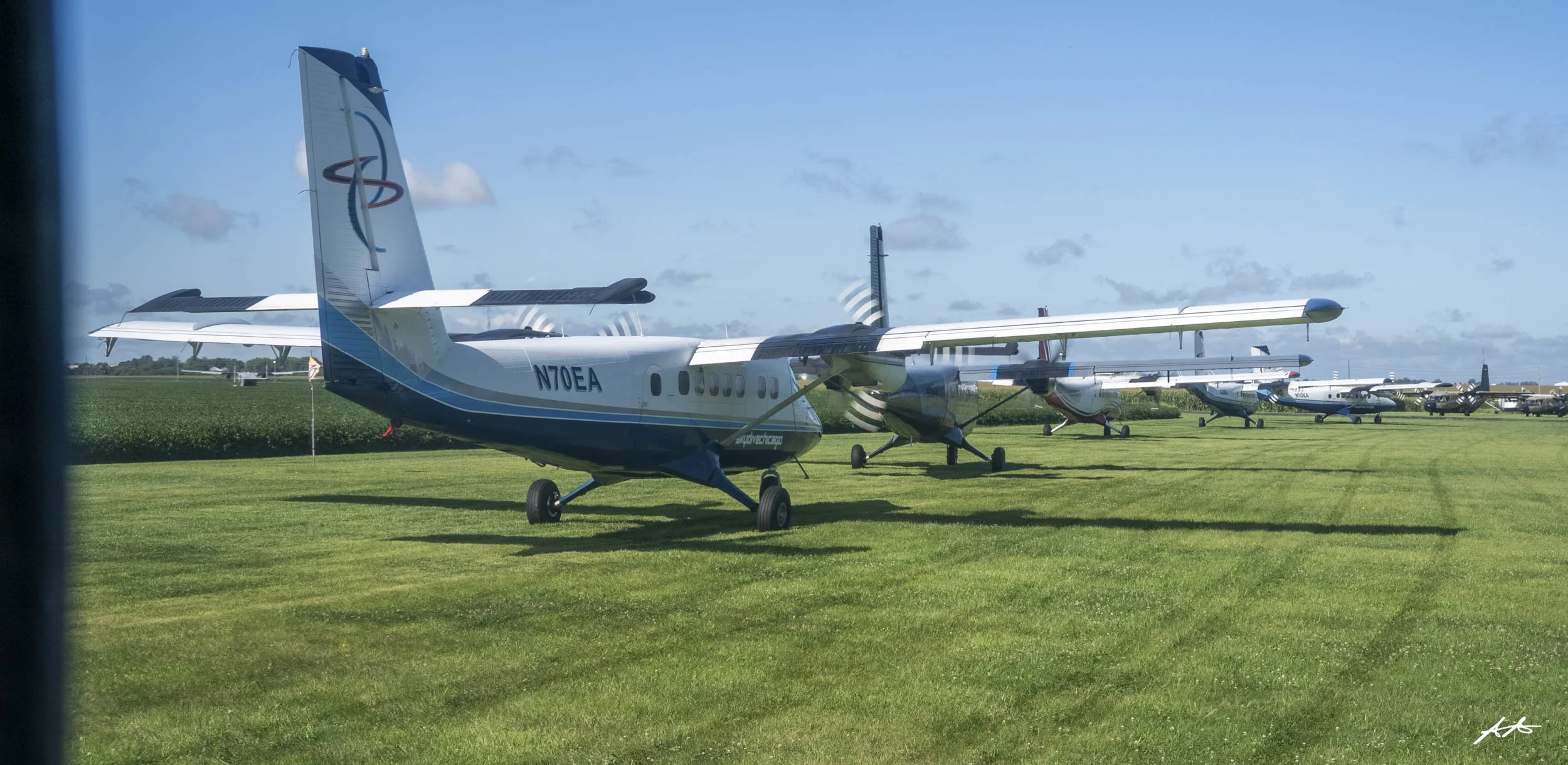 Multiple small airplanes parked on a grassy airfield under a partly cloudy sky.