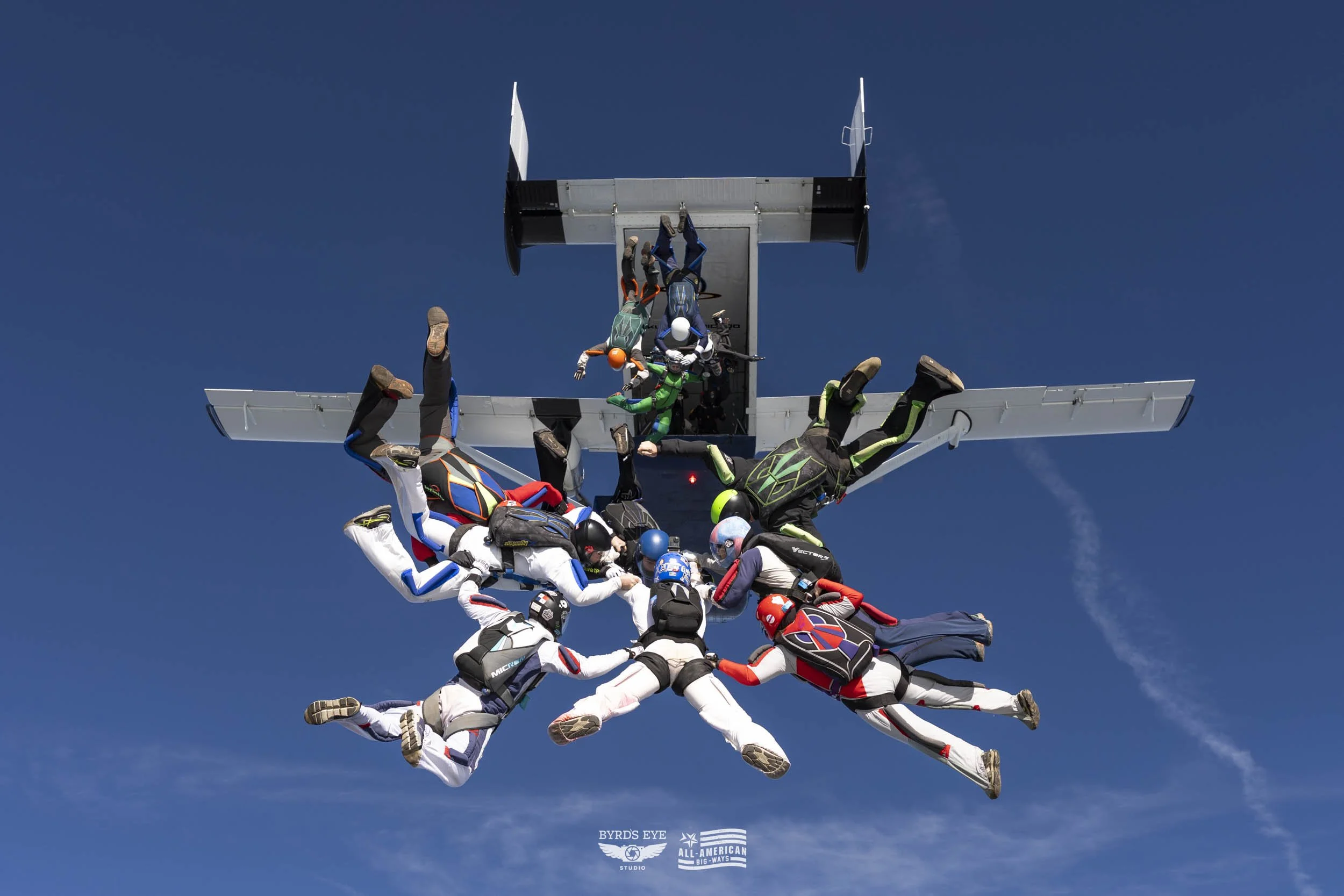 Skydivers holding hands in a circle after jumping from an airplane, with clear blue sky in background.