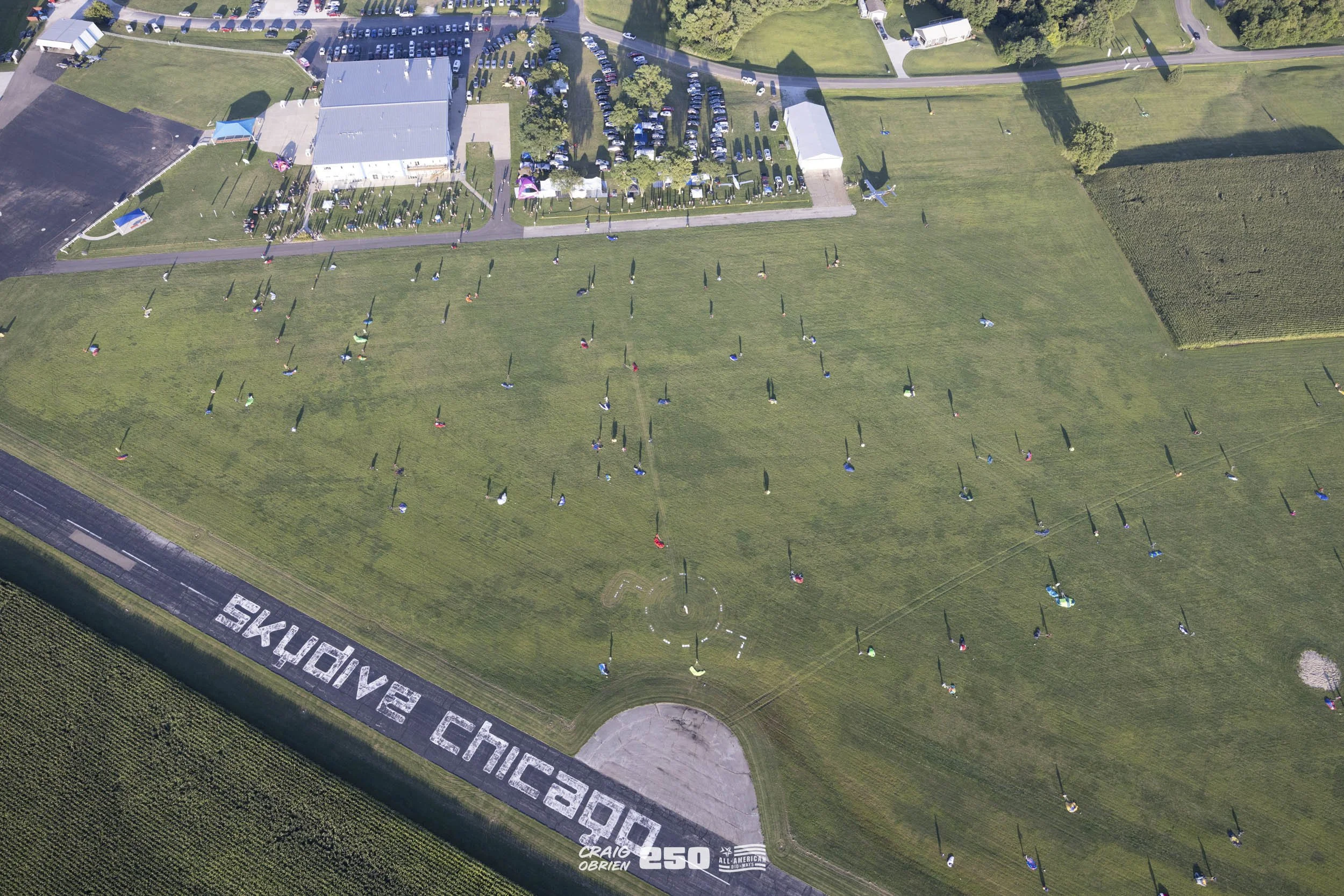 Aerial view of a large cemetery with numerous headstones and a flat green field, with some trees, a building, parking lot, and vehicles in the background. There is a message on the grass that reads 'SAY MY NAME' and some additional text.
