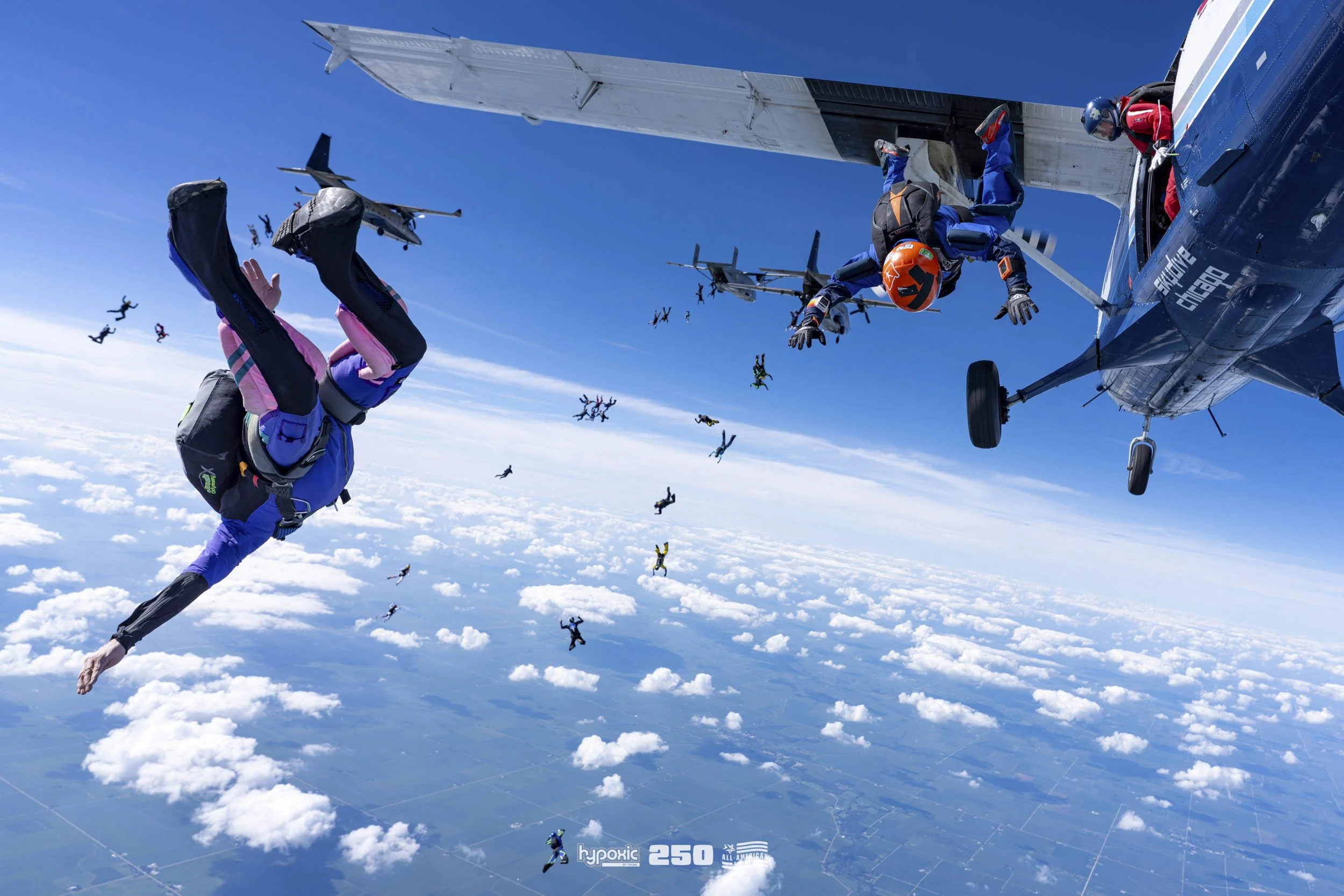Skydivers in free fall with a blue sky and clouds below, some skydivers are falling away from an airplane, others are near the aircraft or mid-air, wearing helmets and jumpsuits.