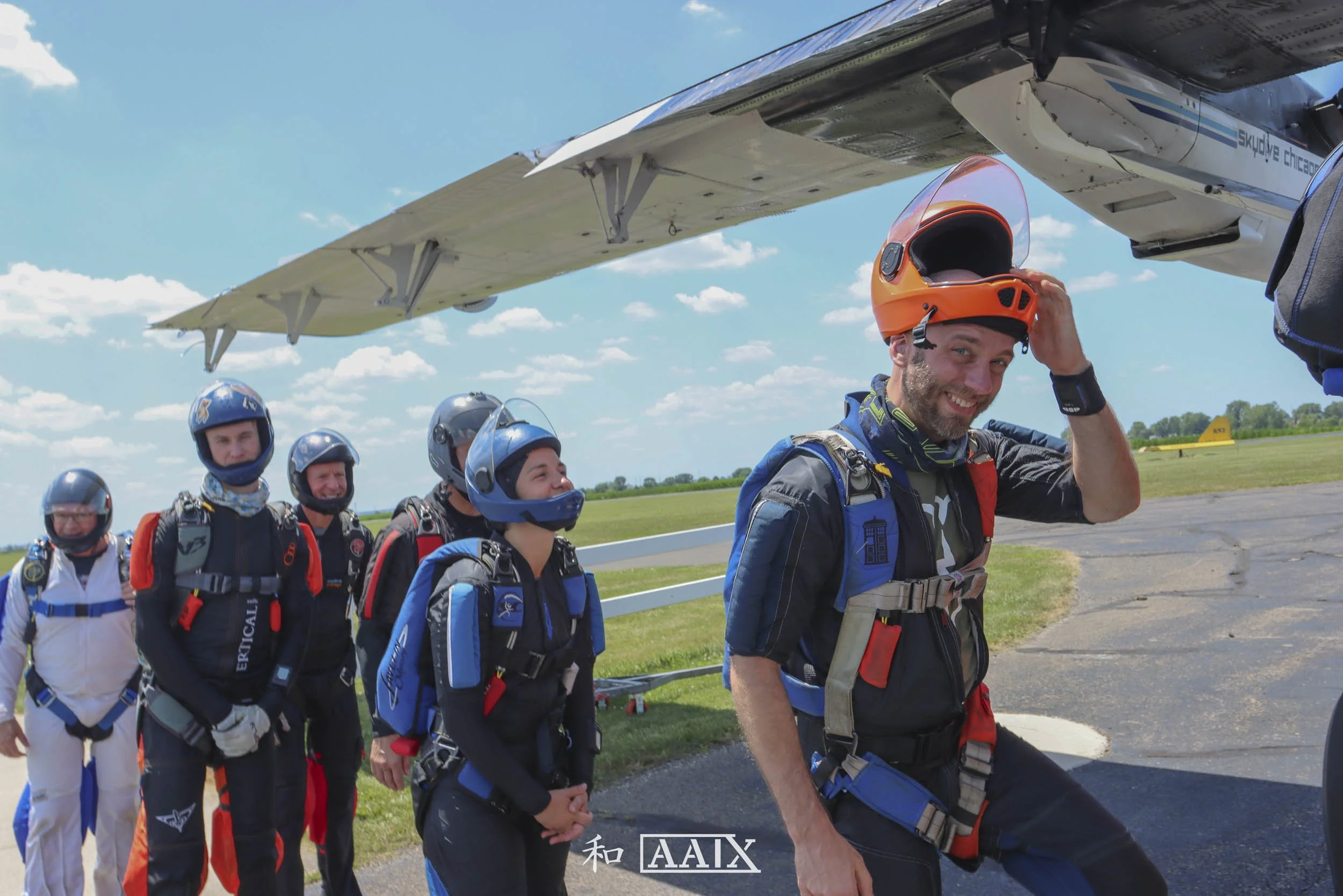 Group of skydivers standing in line on a grassy airfield, preparing for a jump, with one smiling skydiver adjusting his helmet near a plane.