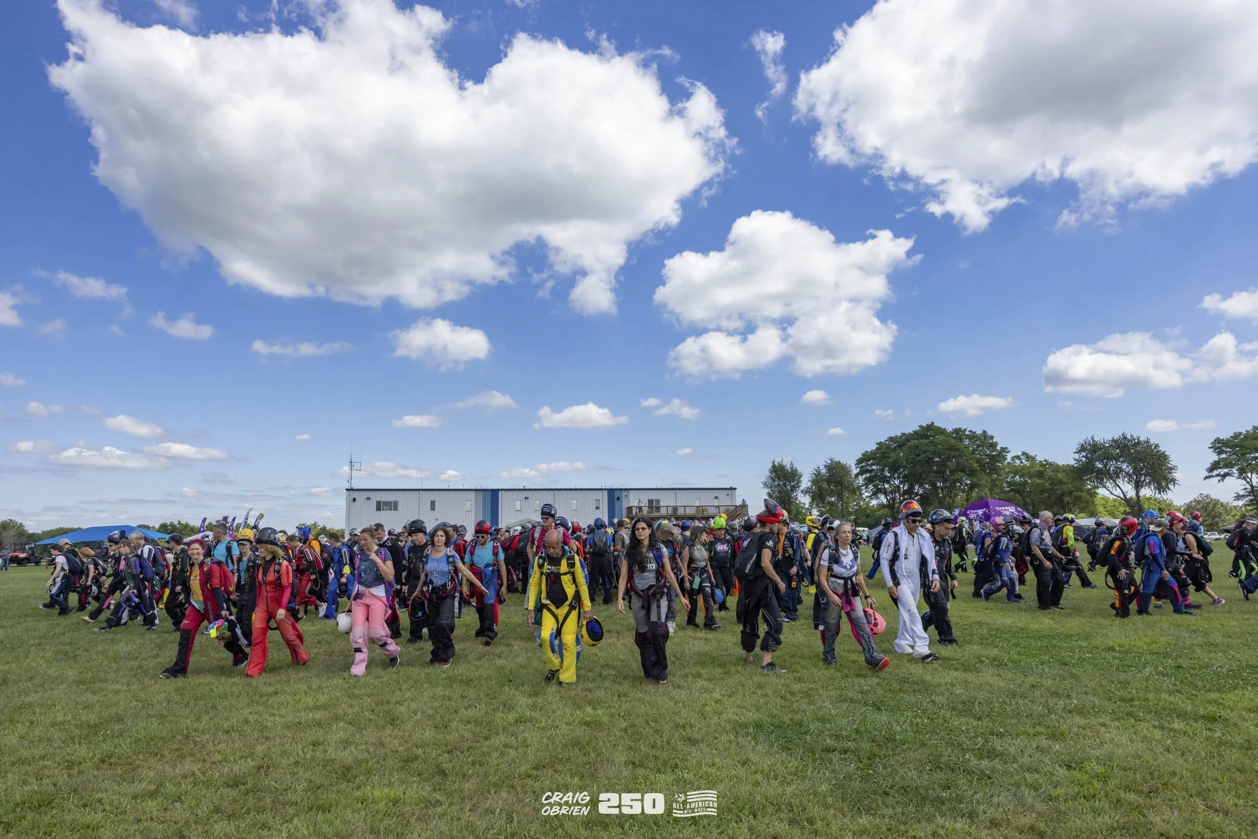 Large group of skydivers in jumpsuits and helmets walking across a grassy field with a blue sky and white clouds above, and a large building in the background.