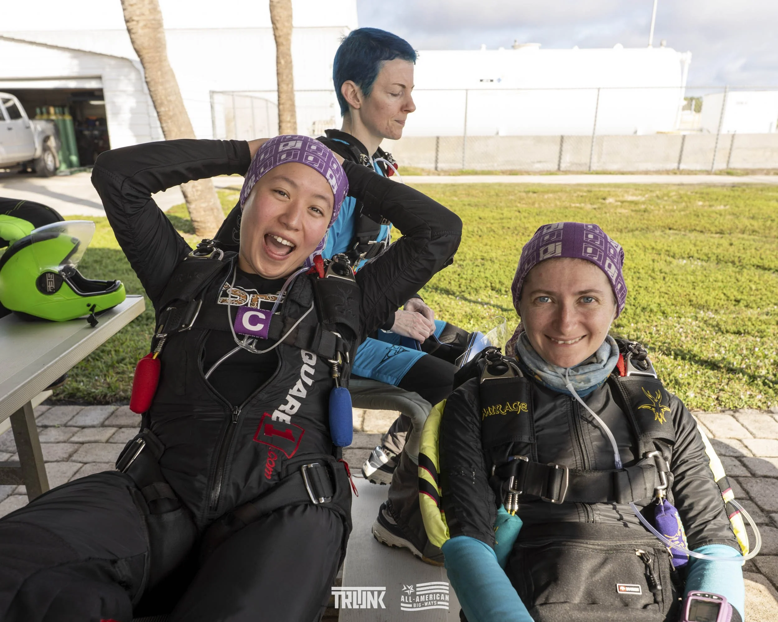 Three women in skydiving suits and head coverings sit on a bench outdoors. One woman is smiling at the camera, the second is behind her looking downward, and the third is in the background adjusting her suit. Skydiving gear, including a green helmet,