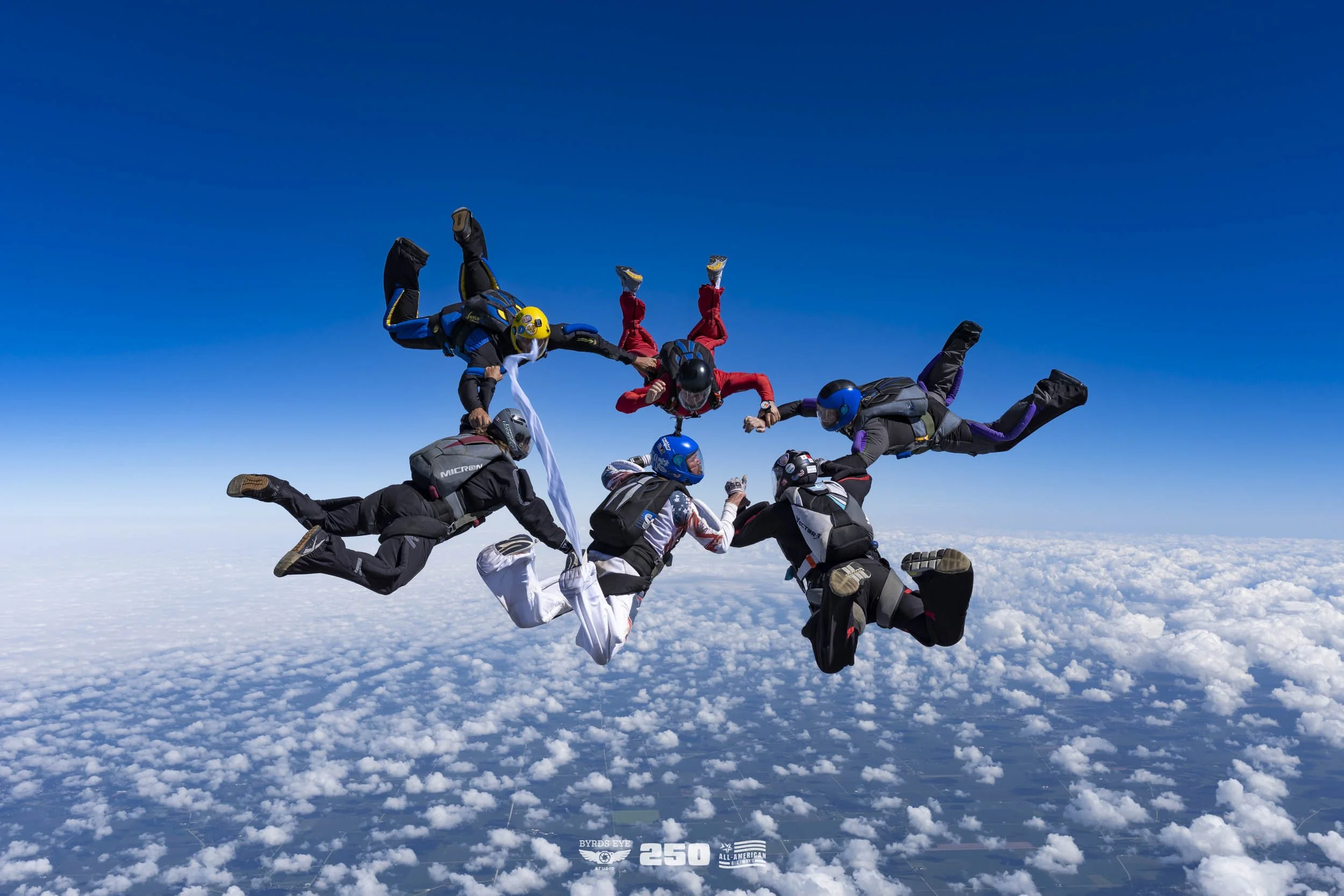 Group of skydivers holding hands in a circle mid-air above clouds with a clear blue sky.