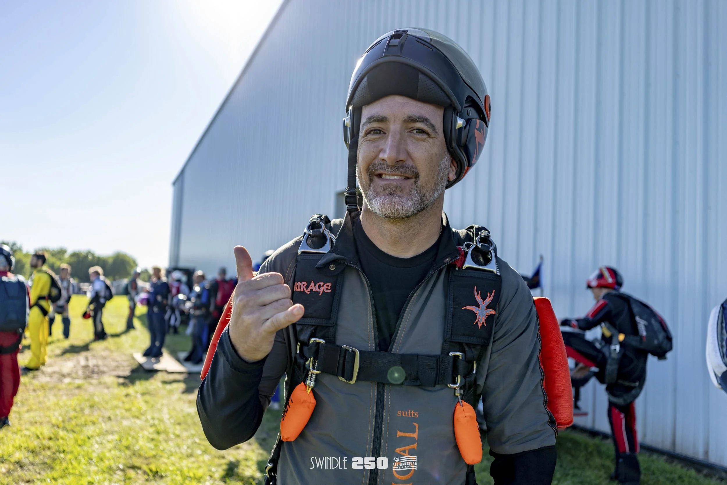 A man in skydiving gear giving a thumbs-up at an outdoor skydiving facility, with other skydivers preparing in the background.