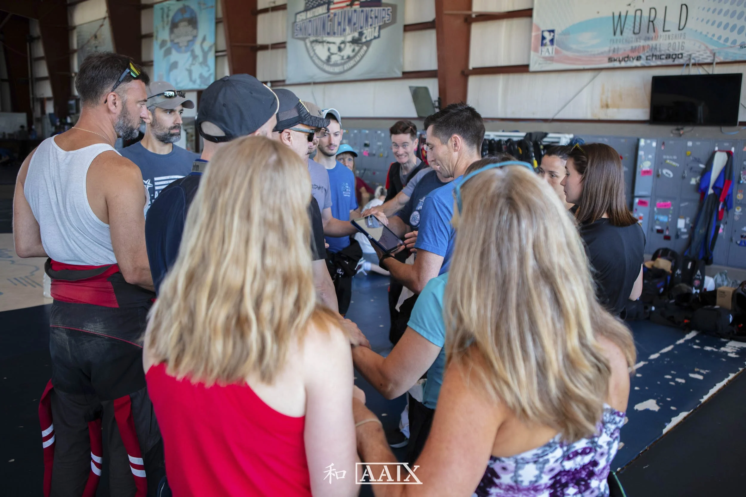 Group of people gathered in an indoor space with banners on the wall, some wearing sunglasses and casual athletic clothing, possibly at a sports event or training session.