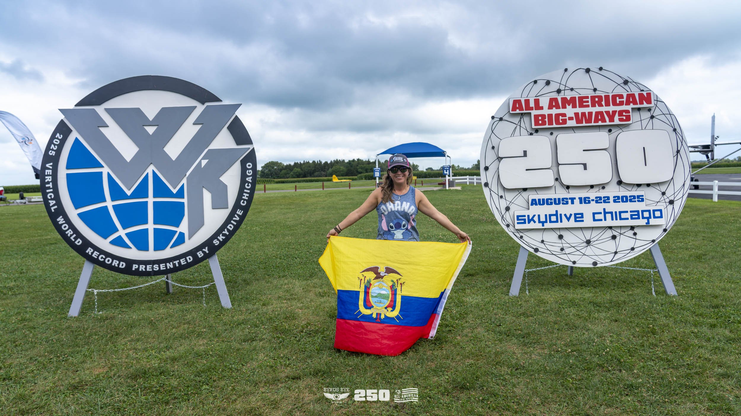 A woman standing outdoors on grassy field holding a Ecuadorian flag, with large signs behind her celebrating a world record for an all-American skydiving event at Skydive Chicago from August 16-22, 2025.