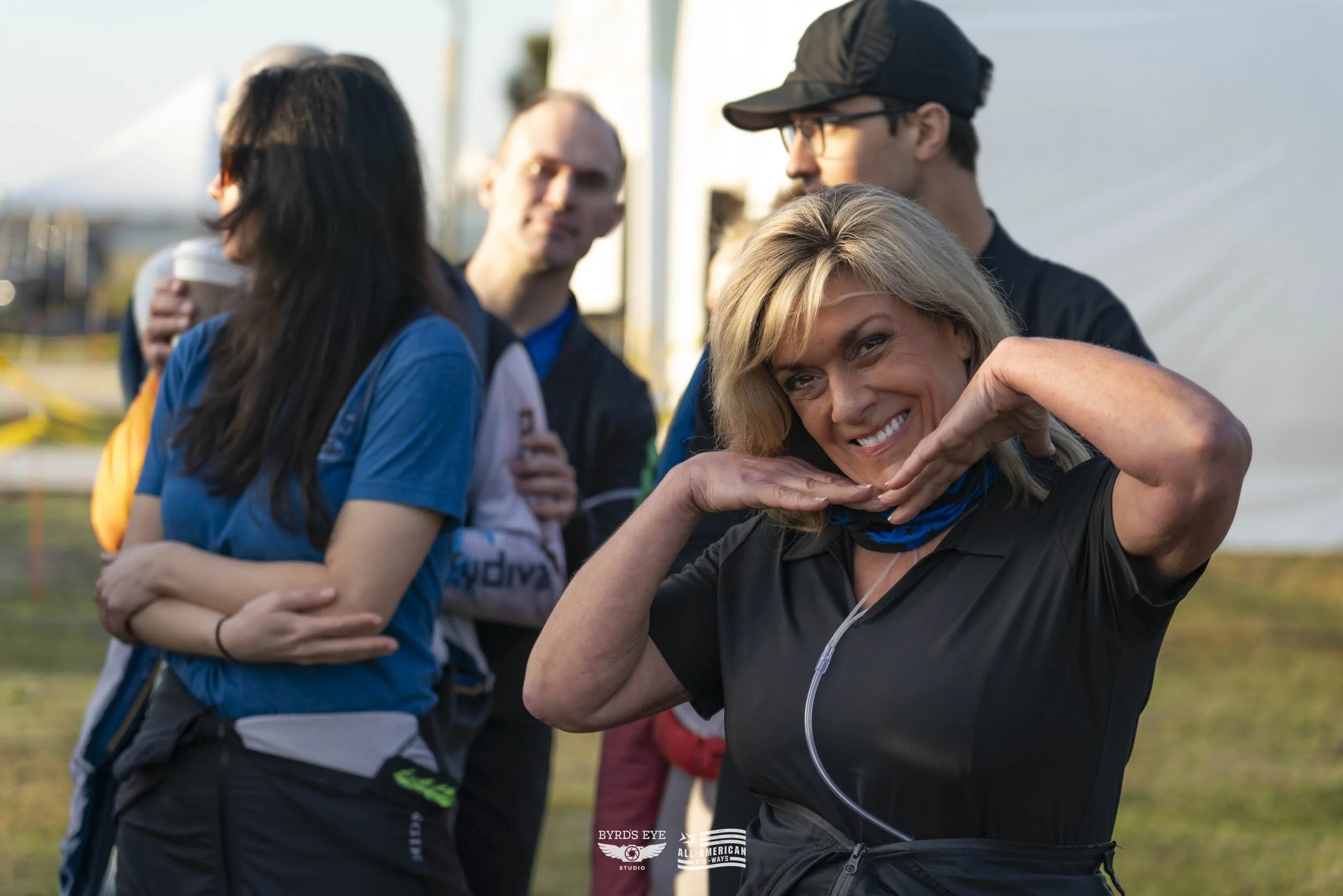 A woman smiling and posing with her hands near her face in a playful manner, with a group of people in the background outdoors during daytime.