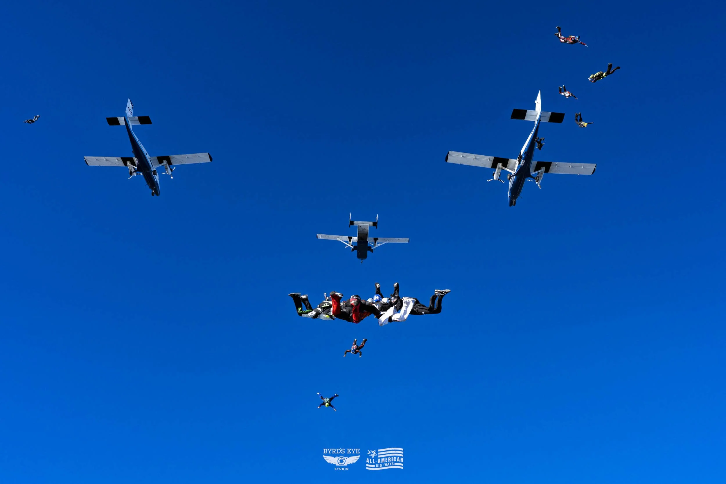 Multiple skydivers free-falling in formation under blue sky with several airplanes flying above.