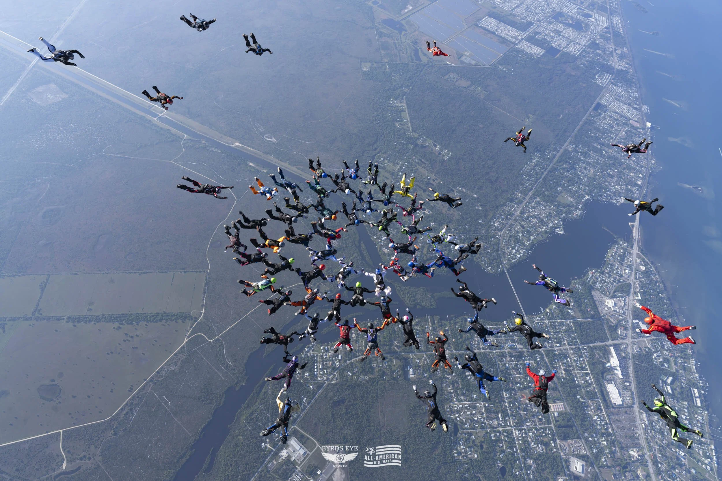 A large group of skydivers in various colored jumpsuits and helmets free-falling over a landscape with rivers, roads, and urban areas.