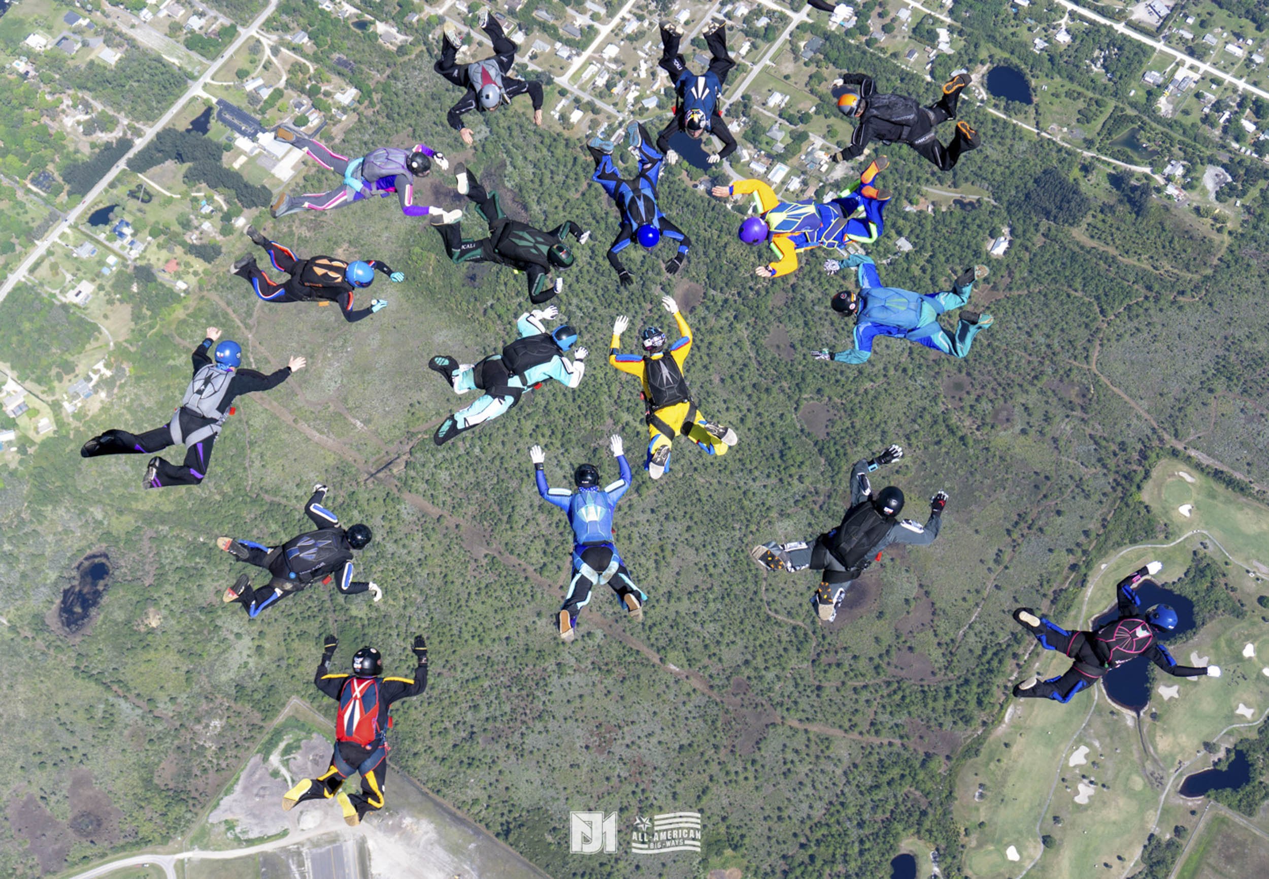Group of skydivers in colorful suits and helmets free-falling over a green landscape with lakes and fields.