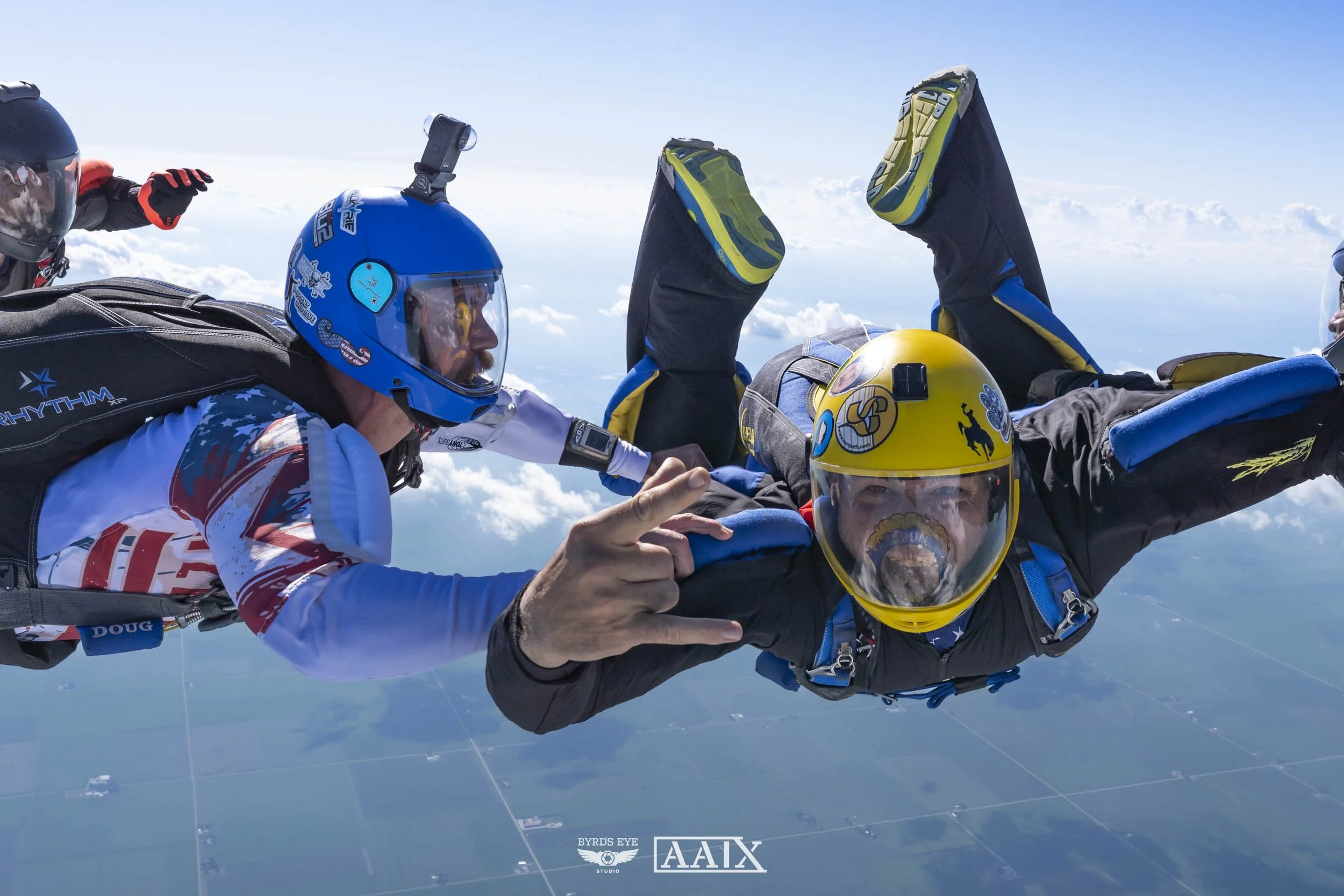 Two skydivers in free fall, wearing helmets and jumpsuits, with clouds and a landscape below, engaging in mid-air formation.