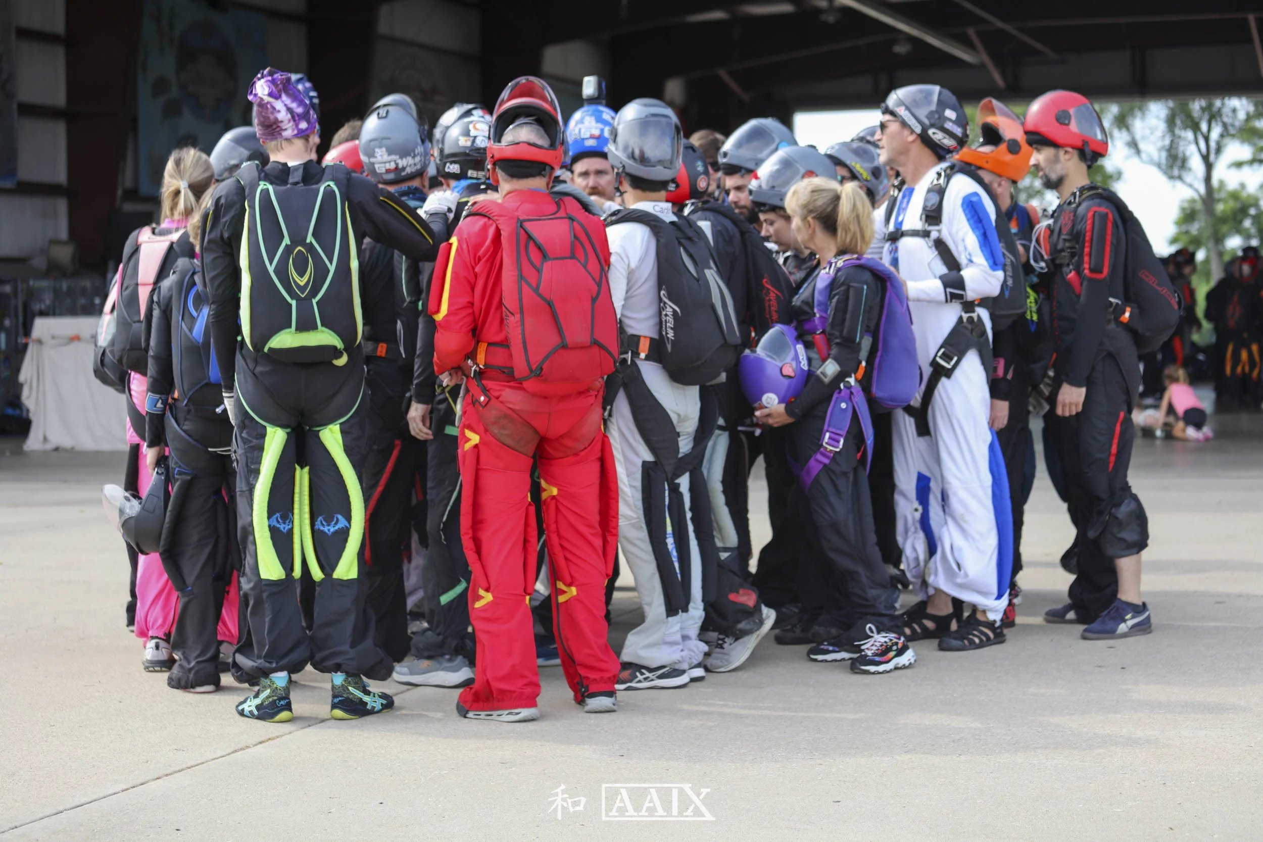 Group of skydivers preparing for a jump, wearing jumpsuits and helmets, gathered in a circle in an indoor hangar.