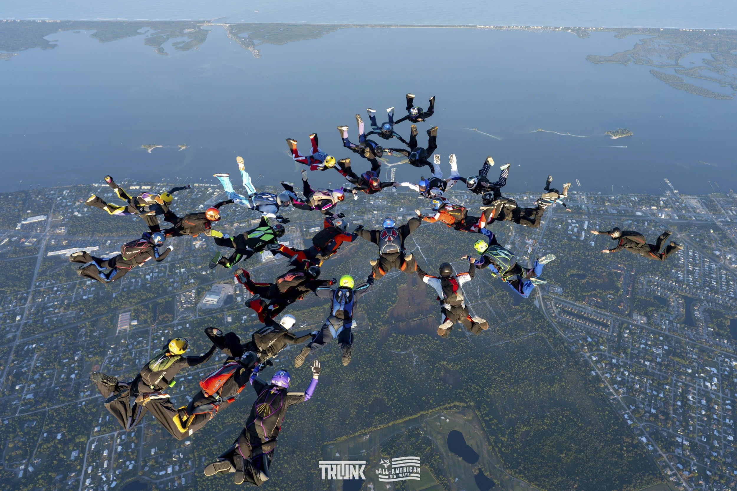 A group of skydivers forming a star shape during free fall over a cityscape with a lake, trees, and buildings below.