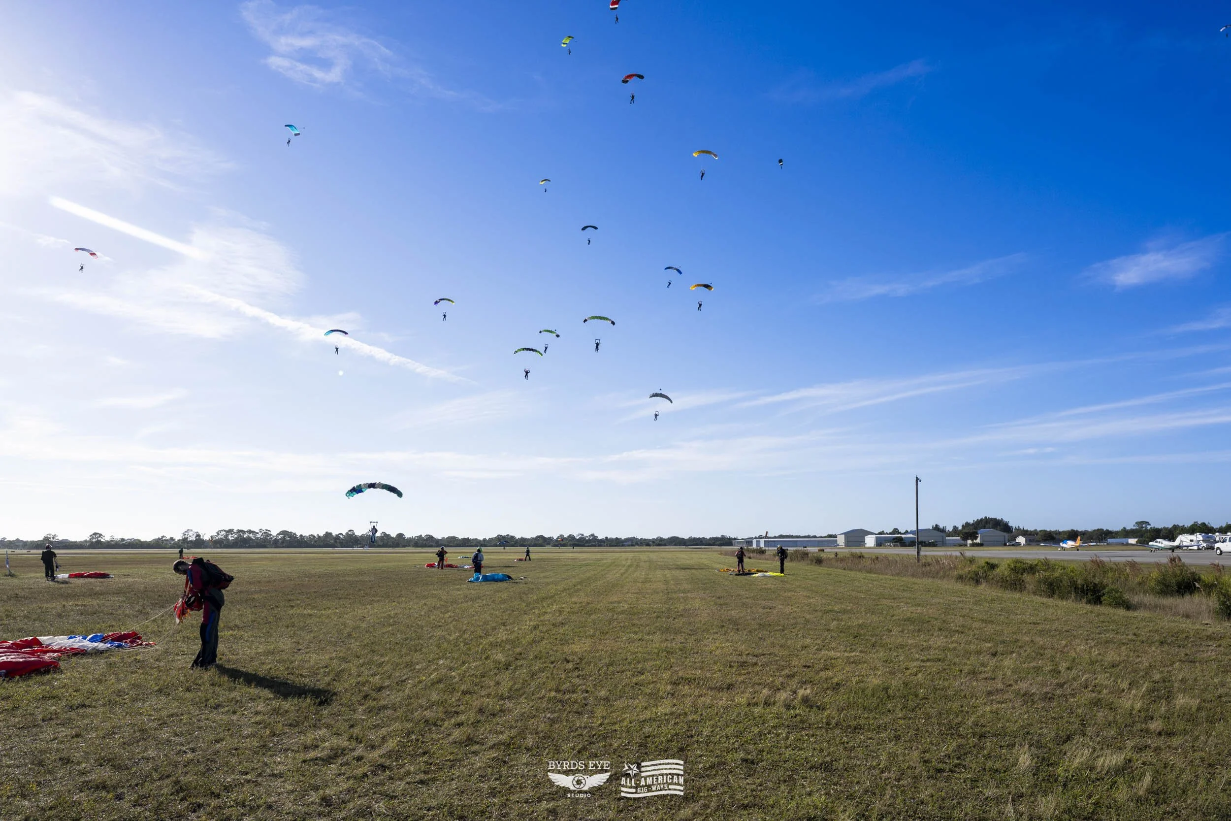 People preparing for paragliding on a grassy field with clear blue sky and several paragliders soaring.