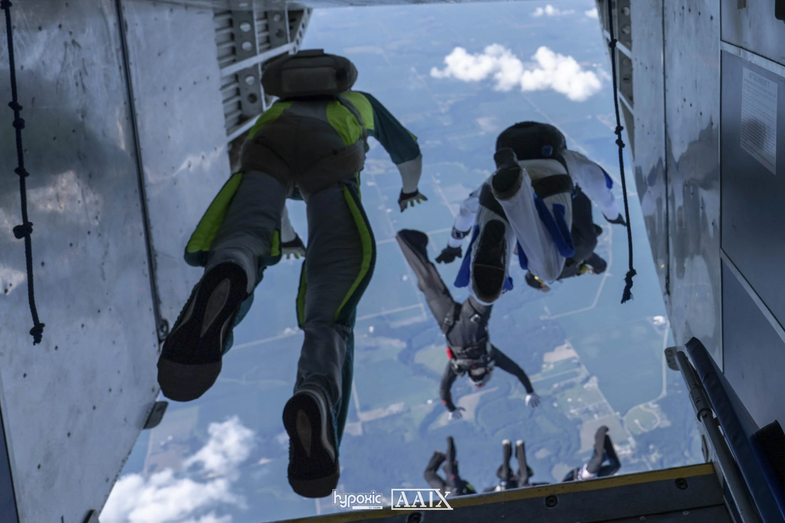 Three skydivers in free fall outside an aircraft, with a view of the earth below and partly cloudy sky.