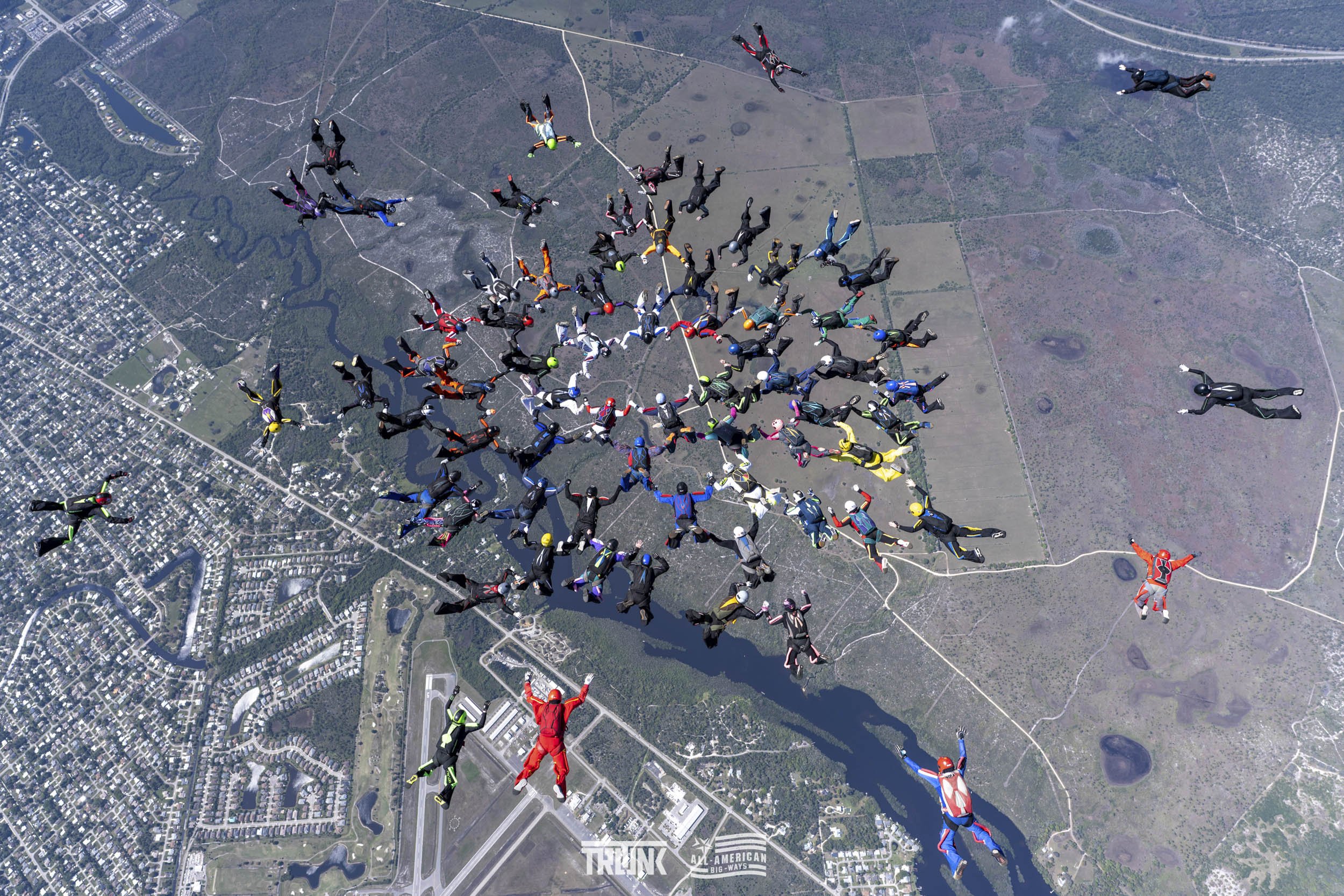 A group of skydivers in colorful jumpsuits and helmets mid-air, forming a formation over a landscape with water, roads, and urban areas.
