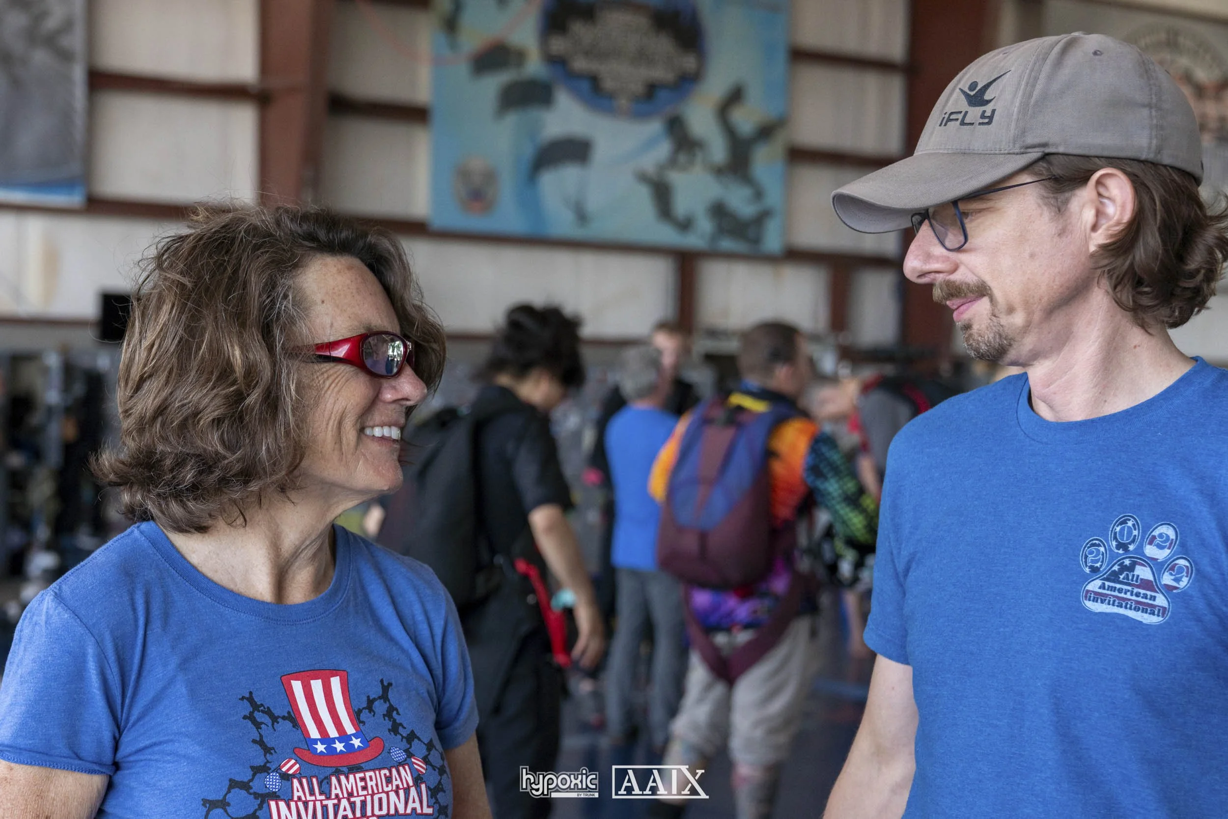A woman and a man are smiling at each other indoors, possibly at a climbing gym, with people and climbing gear in the background. Both are wearing blue shirts with the logo 'All American Invitational.' The woman has curly hair and wears red glasses, 