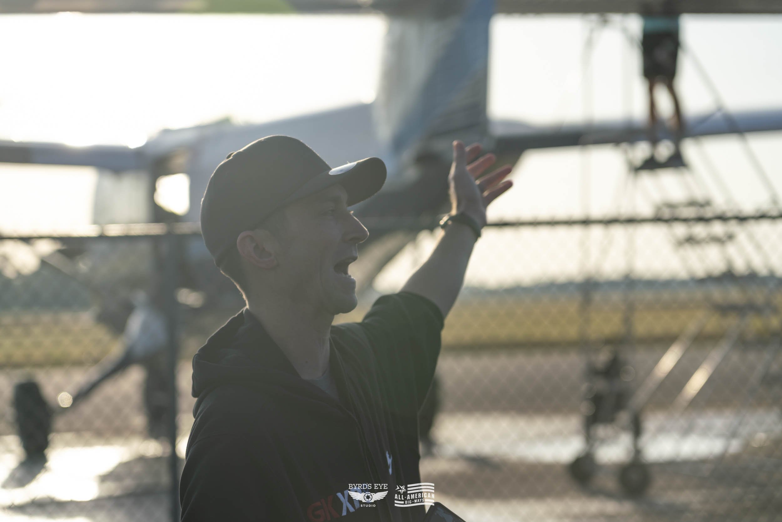 A man wearing a baseball cap and black hoodie gesturing with his arm in front of a plane parked on a tarmac during sunset.