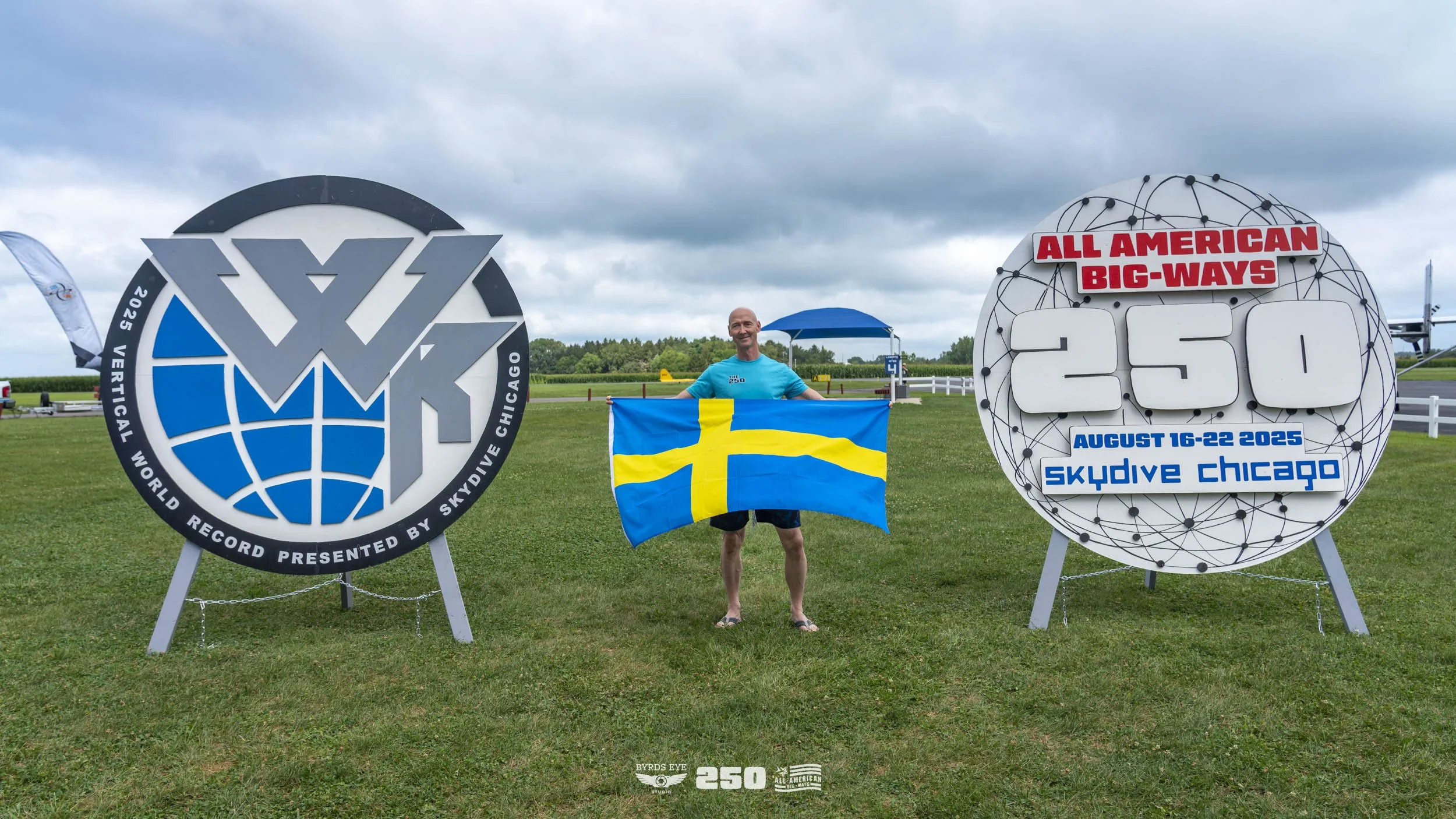 Man standing on grass holding a Swedish flag at Skydiving Chicago, celebrating a world record for 250 skydivers on August 16-22, 2025. Large signs display the event details and record achievement.