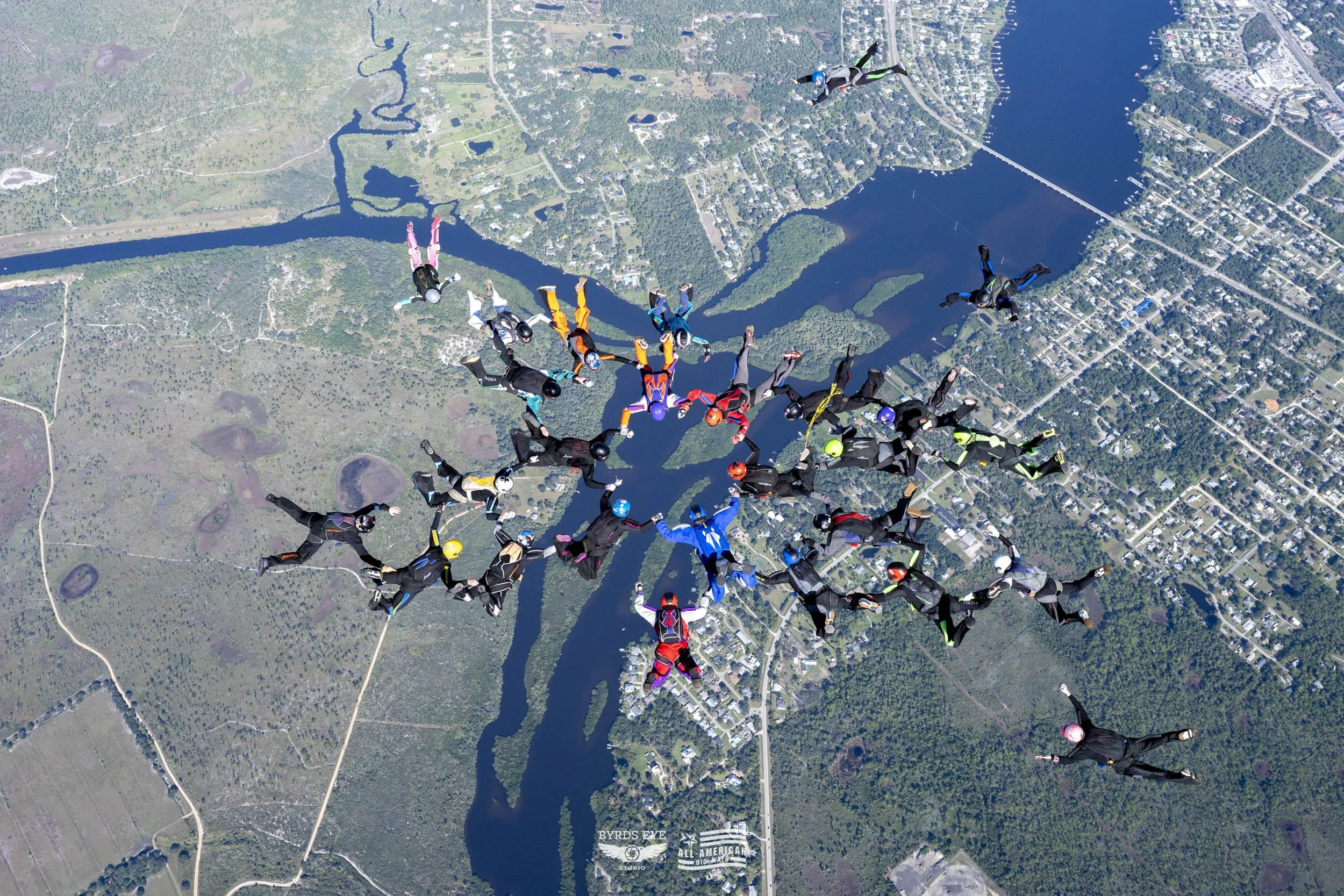 Group of skydivers forming a heart shape while mid-air over a landscape with rivers, forests, and a town.