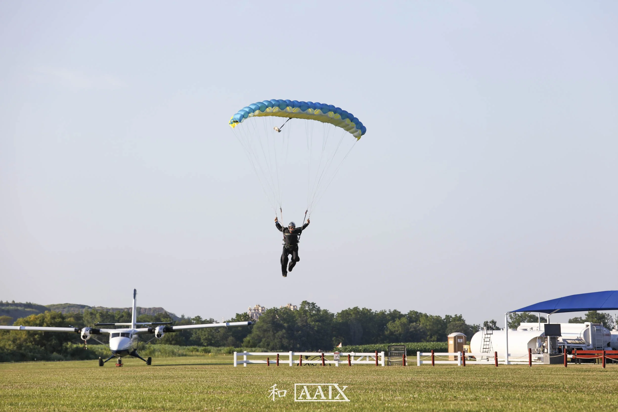 A person skydiving with a parachute, descending towards a small aircraft on the ground in an open field. The sky is clear and blue, and there are trees and structures in the background.