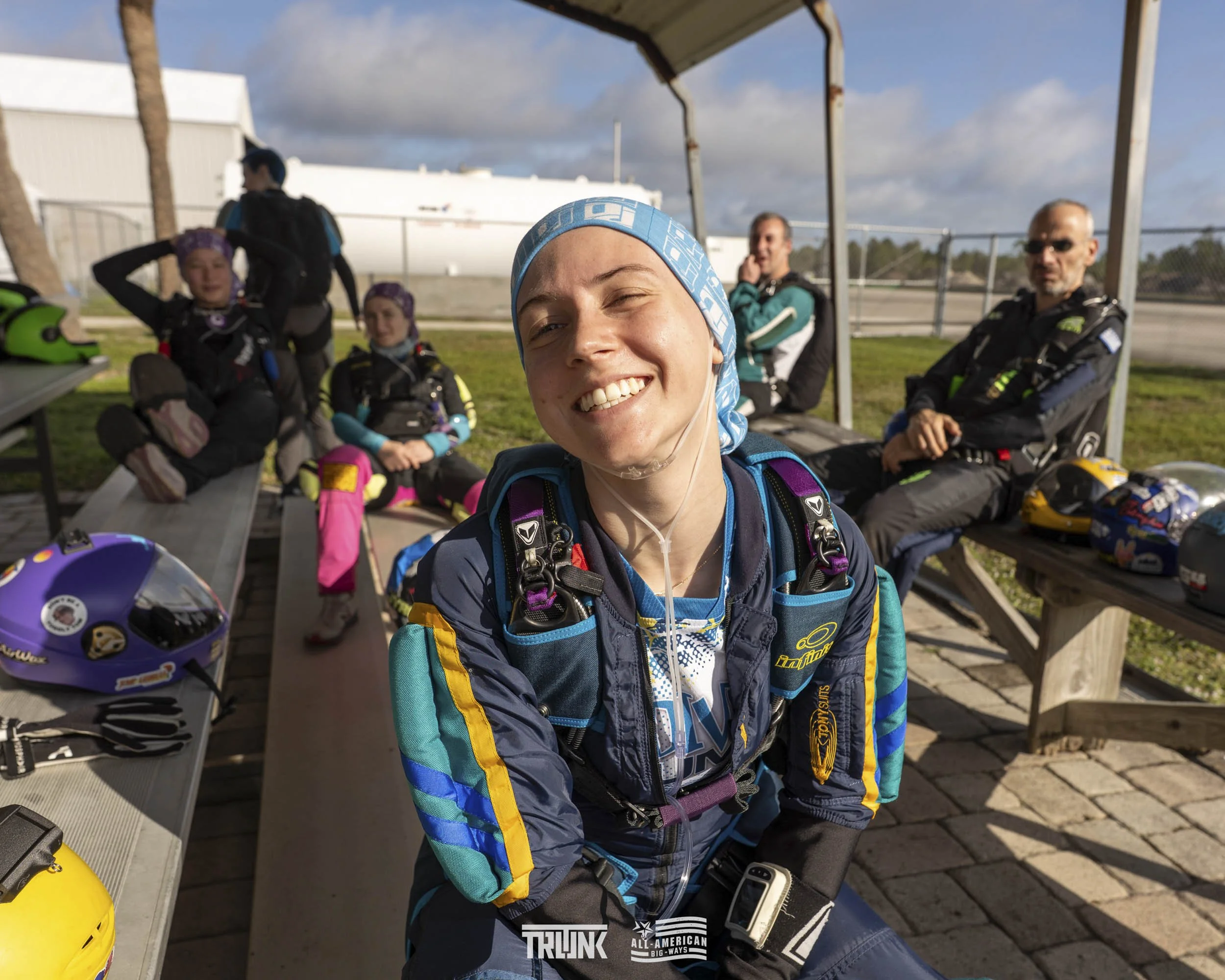 A group of skydivers sitting on benches at a skydiving facility. One woman in the foreground is smiling and looking at the camera, wearing a blue headscarf and skydiving gear. Other people in the background are also dressed in skydiving gear, with he