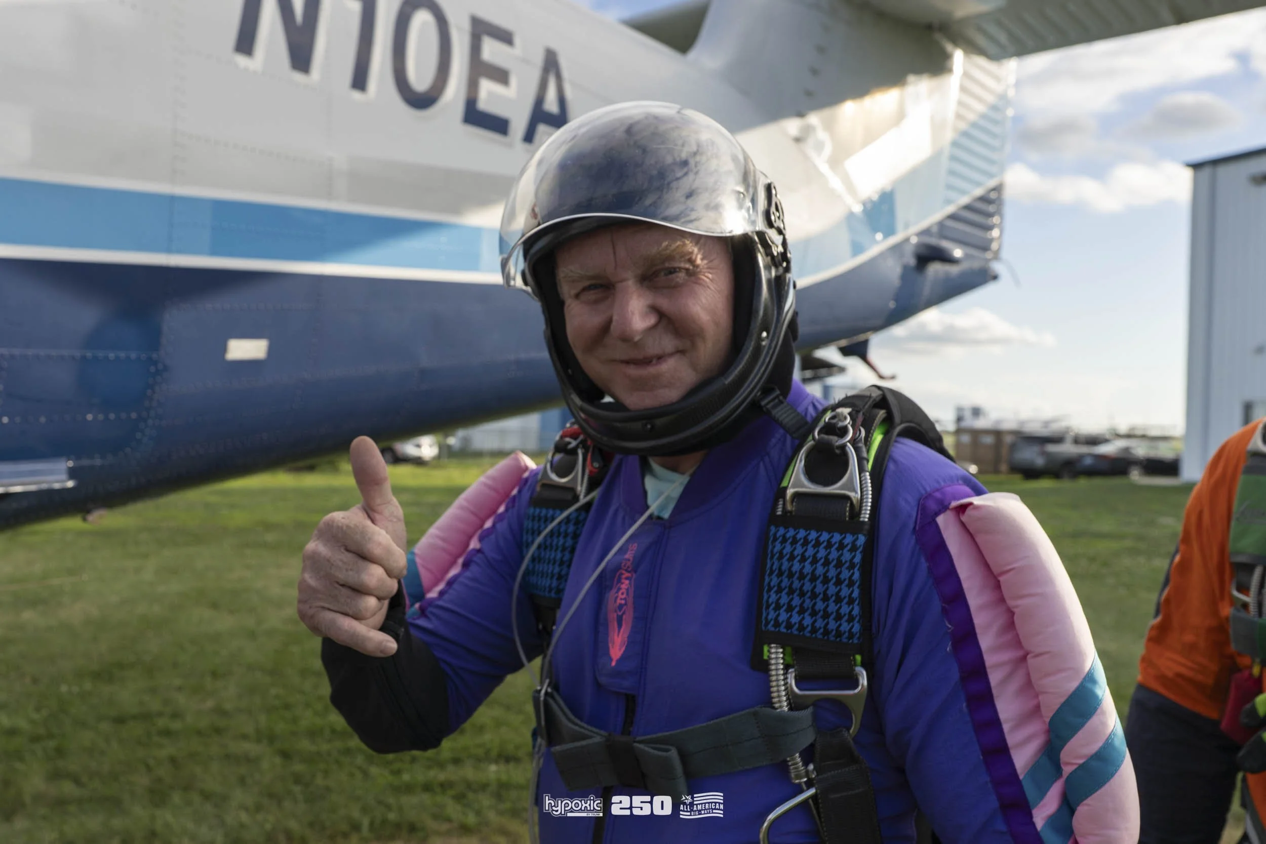 A skydiver in a purple jumpsuit with pink and blue accents giving a thumbs up and smiling, standing on the grass with a plane behind him.