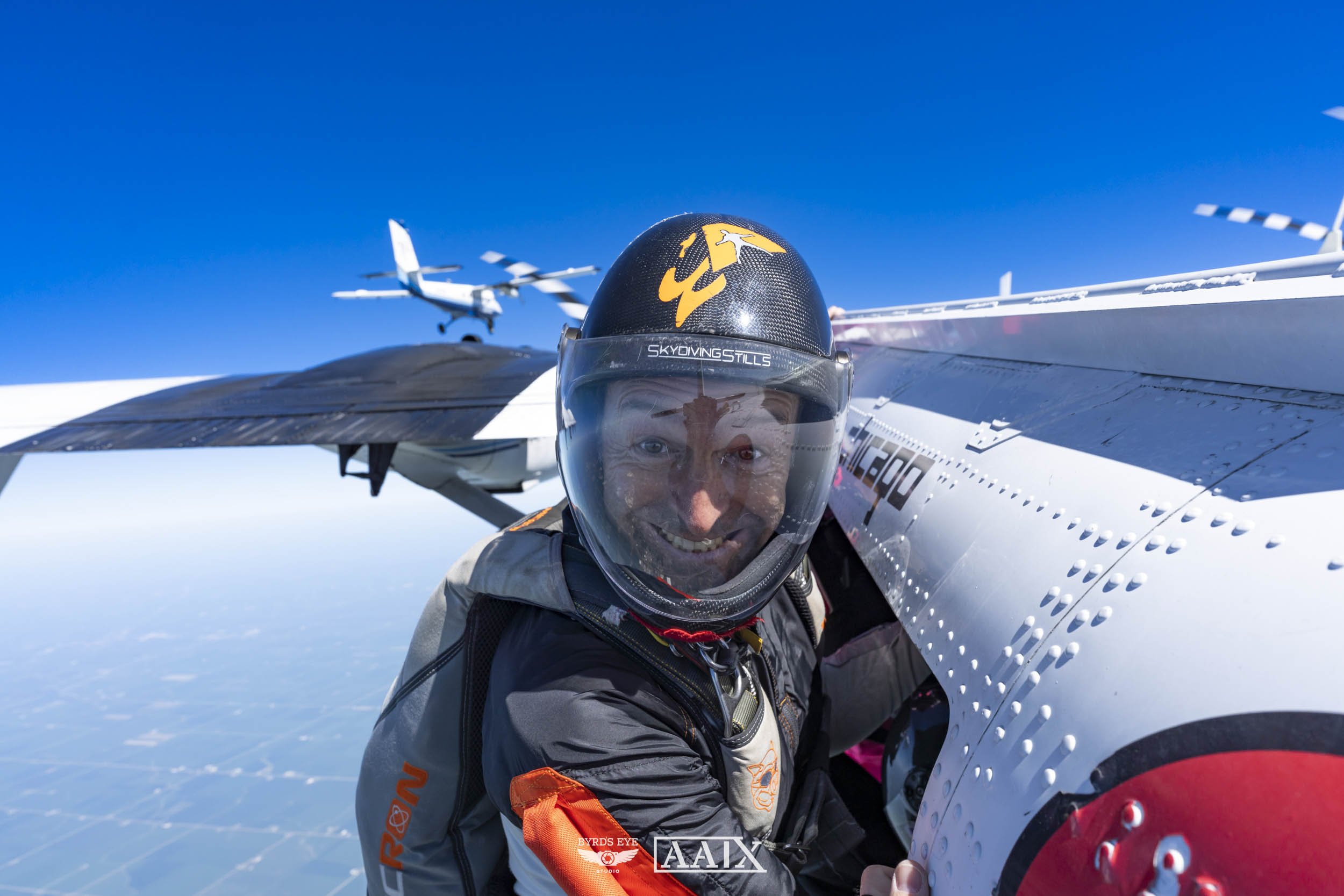 A man in a space suit and helmet mid-air outside a spacecraft, with a plane and sky in the background.