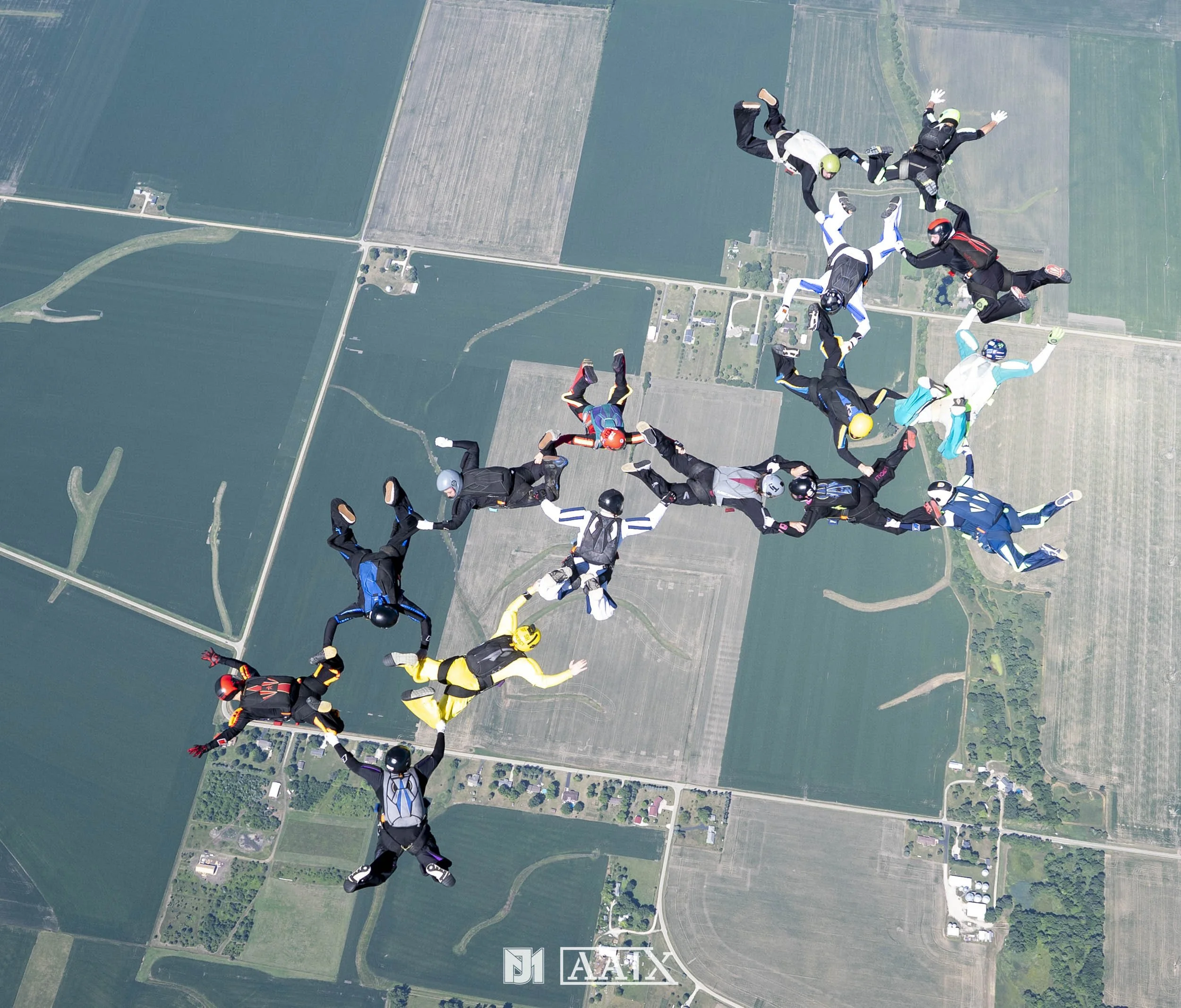 A group of skydivers forming a circle while free-falling over a patchwork of farmland.