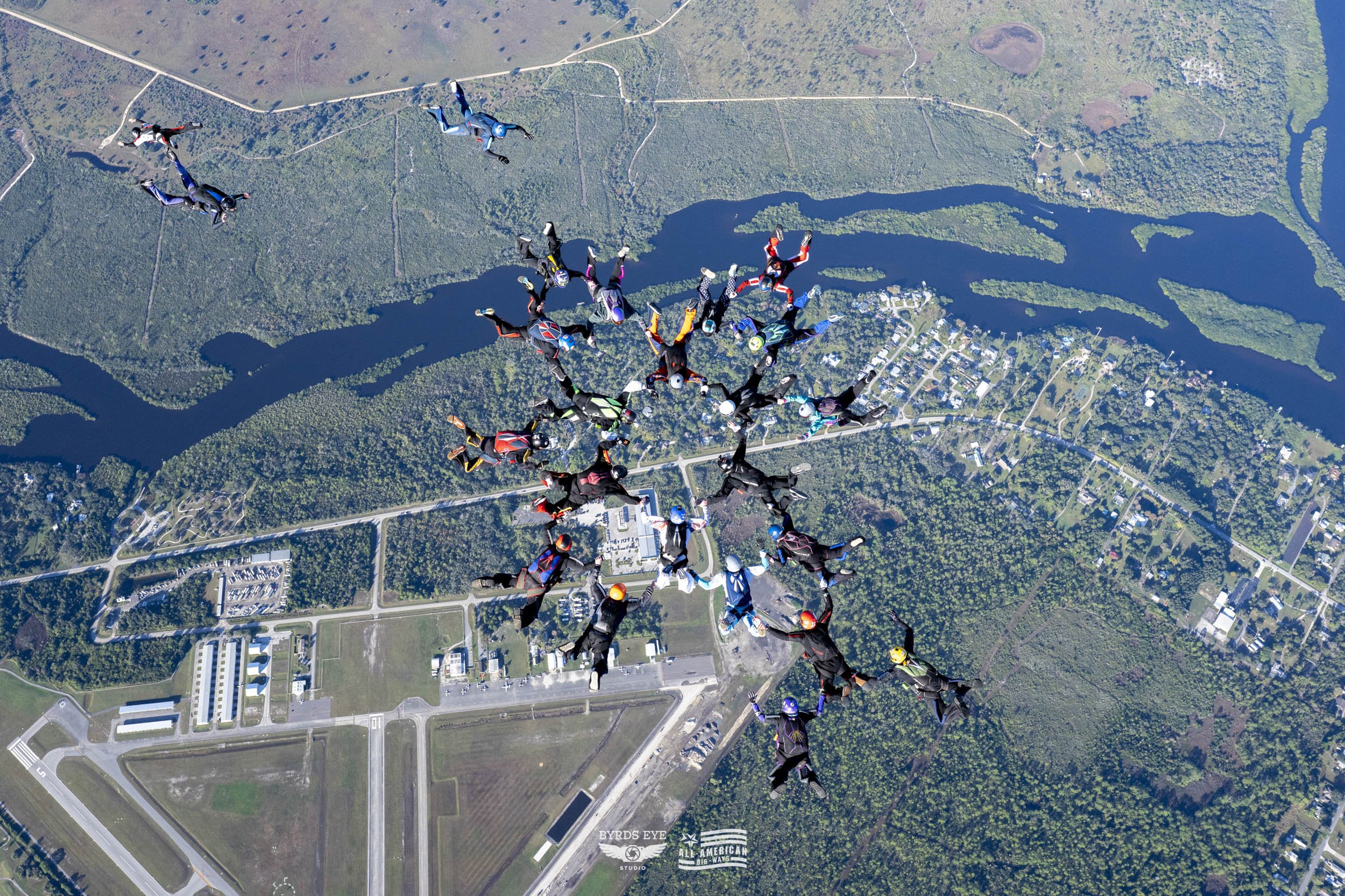 A group of 25 skydivers forming a circle in mid-air over a landscape with a river, trees, and roads below.