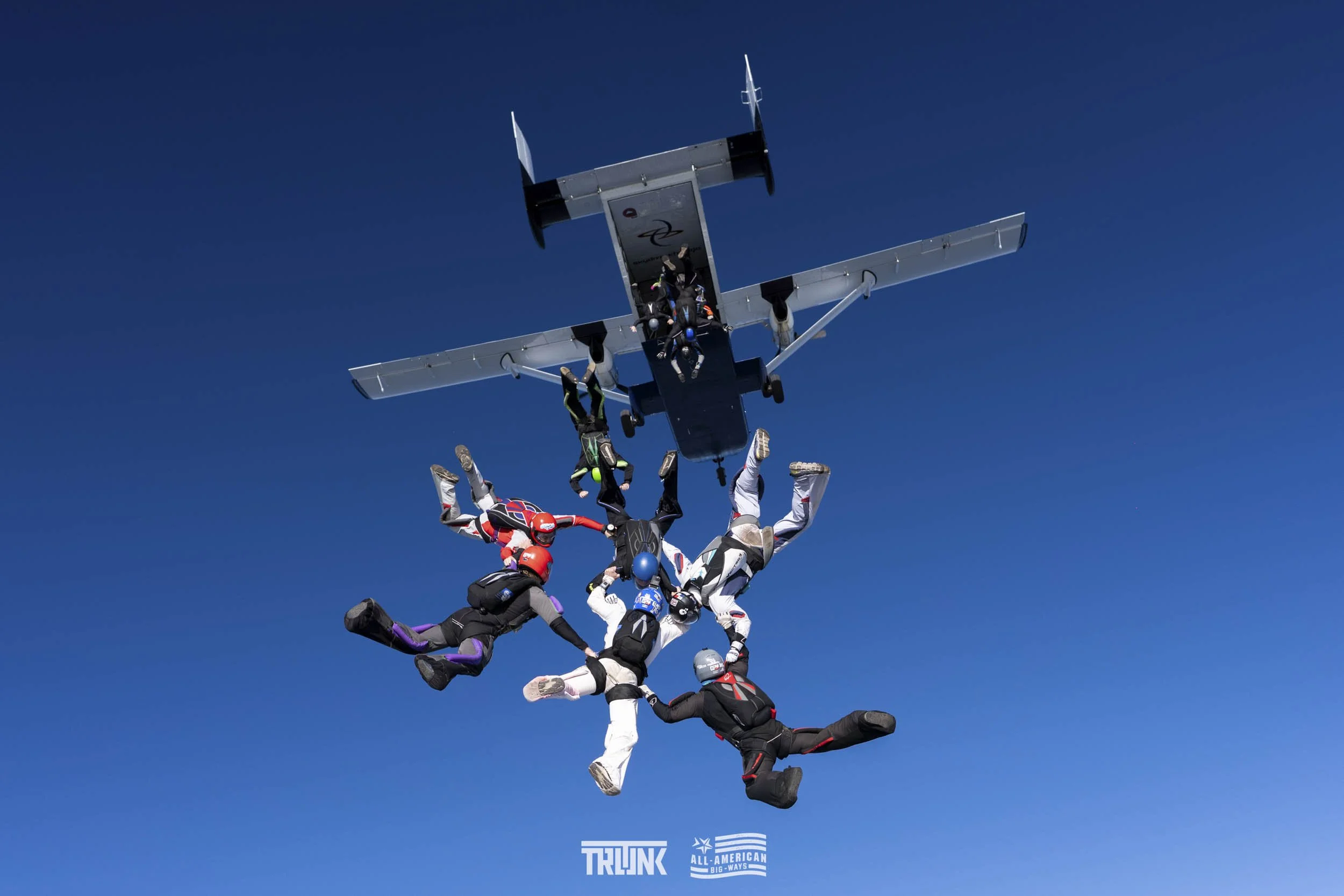 Skydivers performing free-fall formations near an aircraft against a clear blue sky.