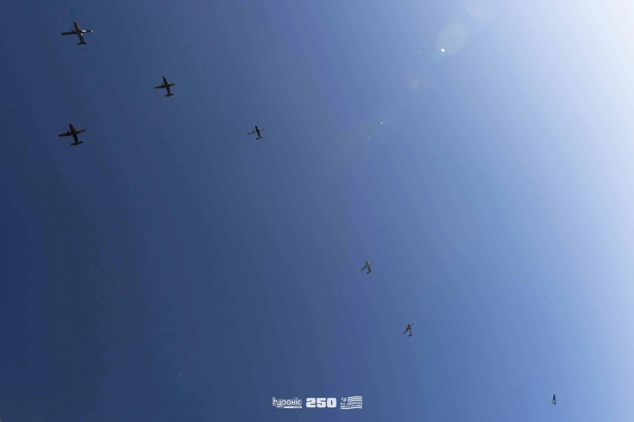A formation of six airplanes flying in a clear blue sky, with the sun shining brightly on the right side of the image.