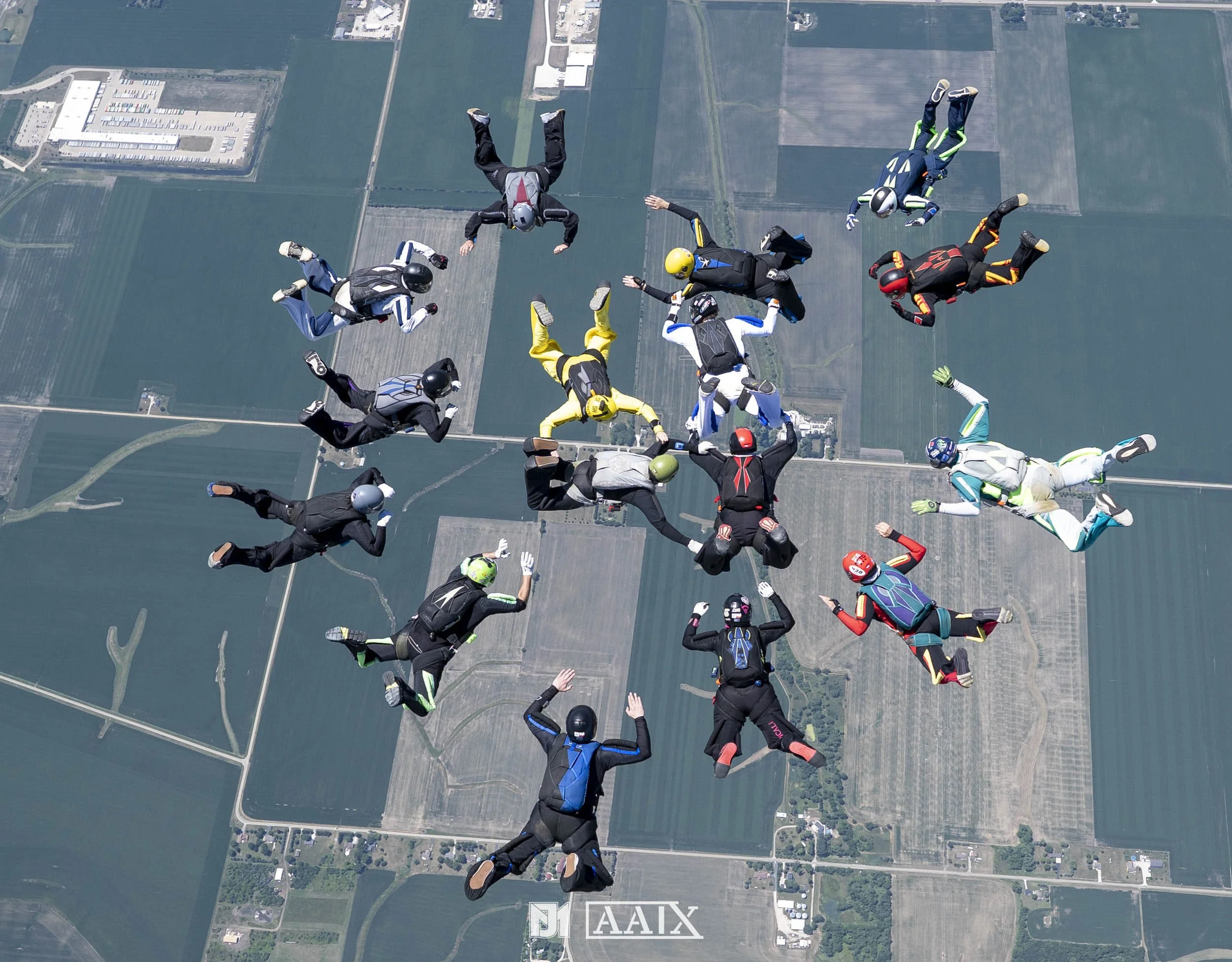 A group of skydivers in colorful jumpsuits and helmets forming a circle while skydiving above a farmland landscape.