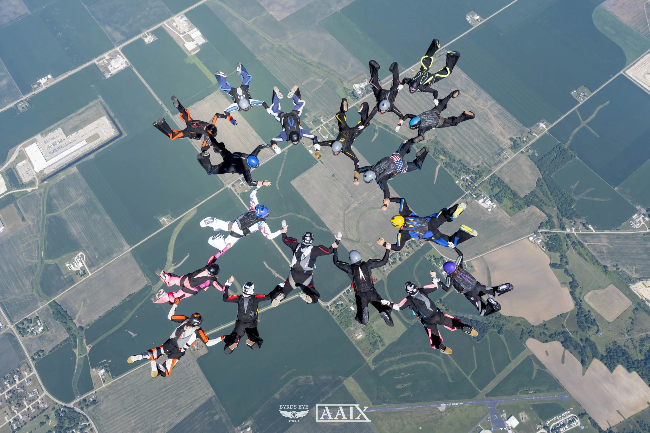 Group of skydivers free-falling in formation above farmland, holding hands in a circle with a landscape of fields below.