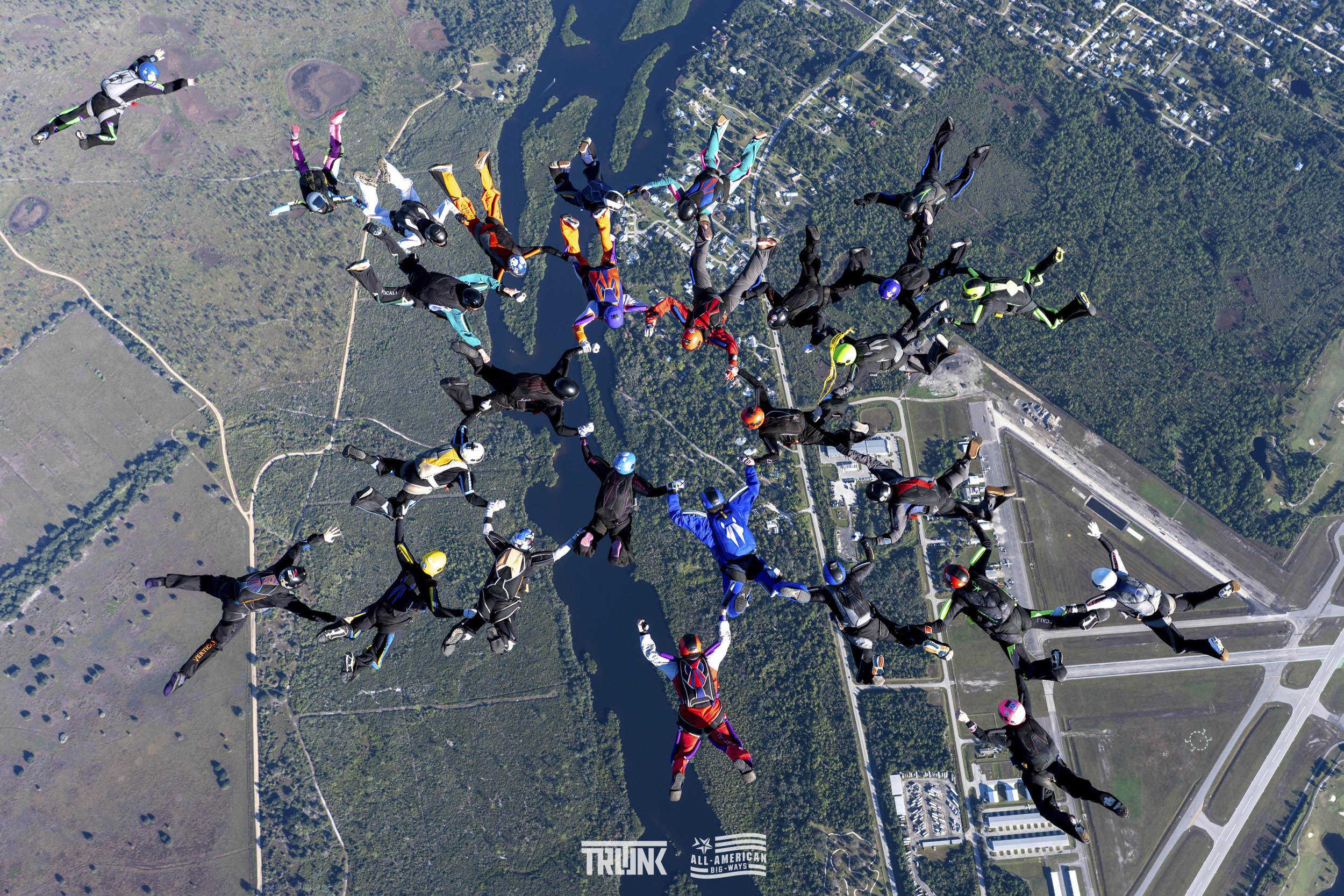 Group of skydivers forming a heart shape while free-flying above a landscape with water, forests, and buildings.