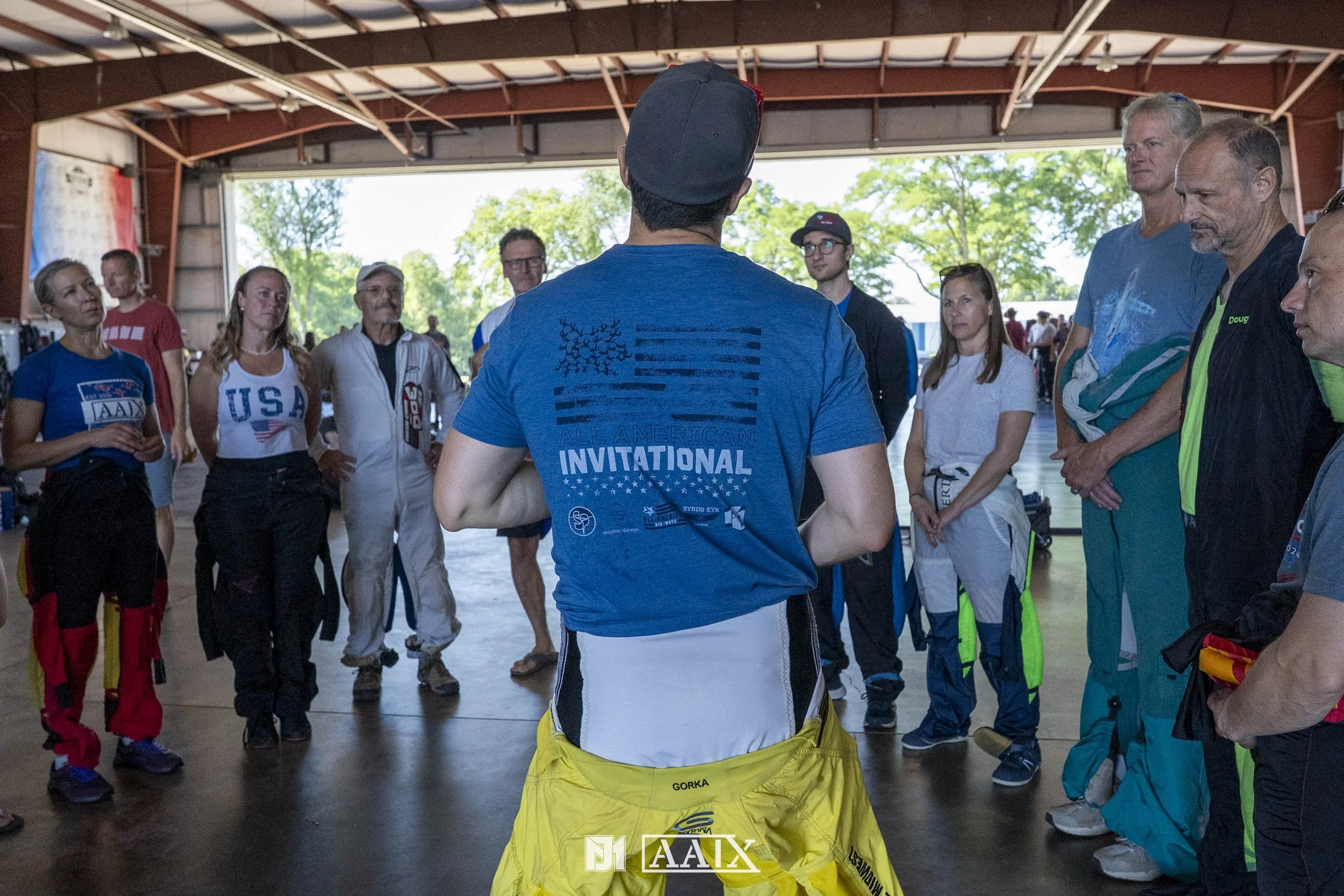 A group of people gathered inside a hangar, listening to a person in a blue T-shirt with the words 'All American Invitational' on the back, who appears to be giving instructions or a talk. The people are diverse in age and are dressed in sports or ca
