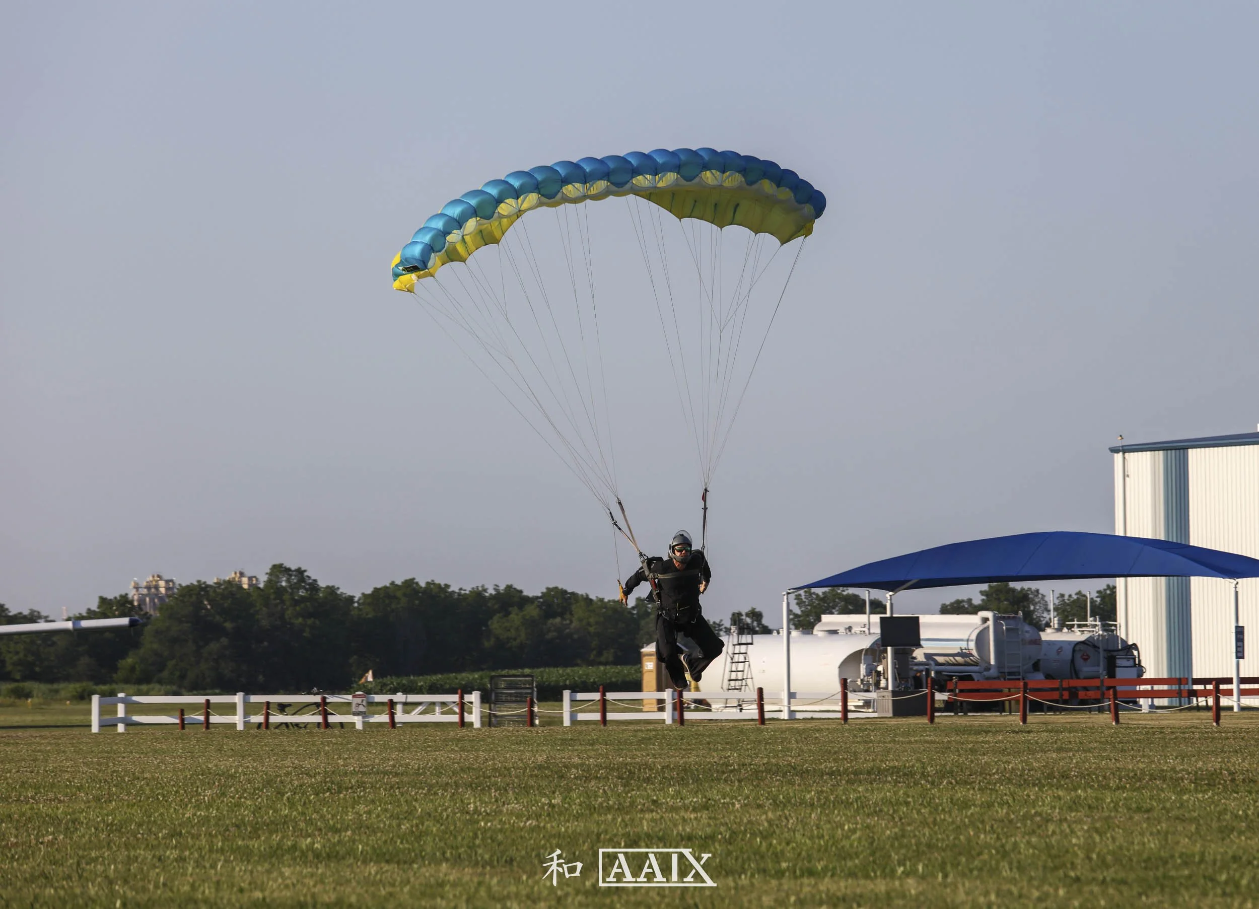 A person is skydiving with a blue and yellow parachute, just above the ground in an open field.