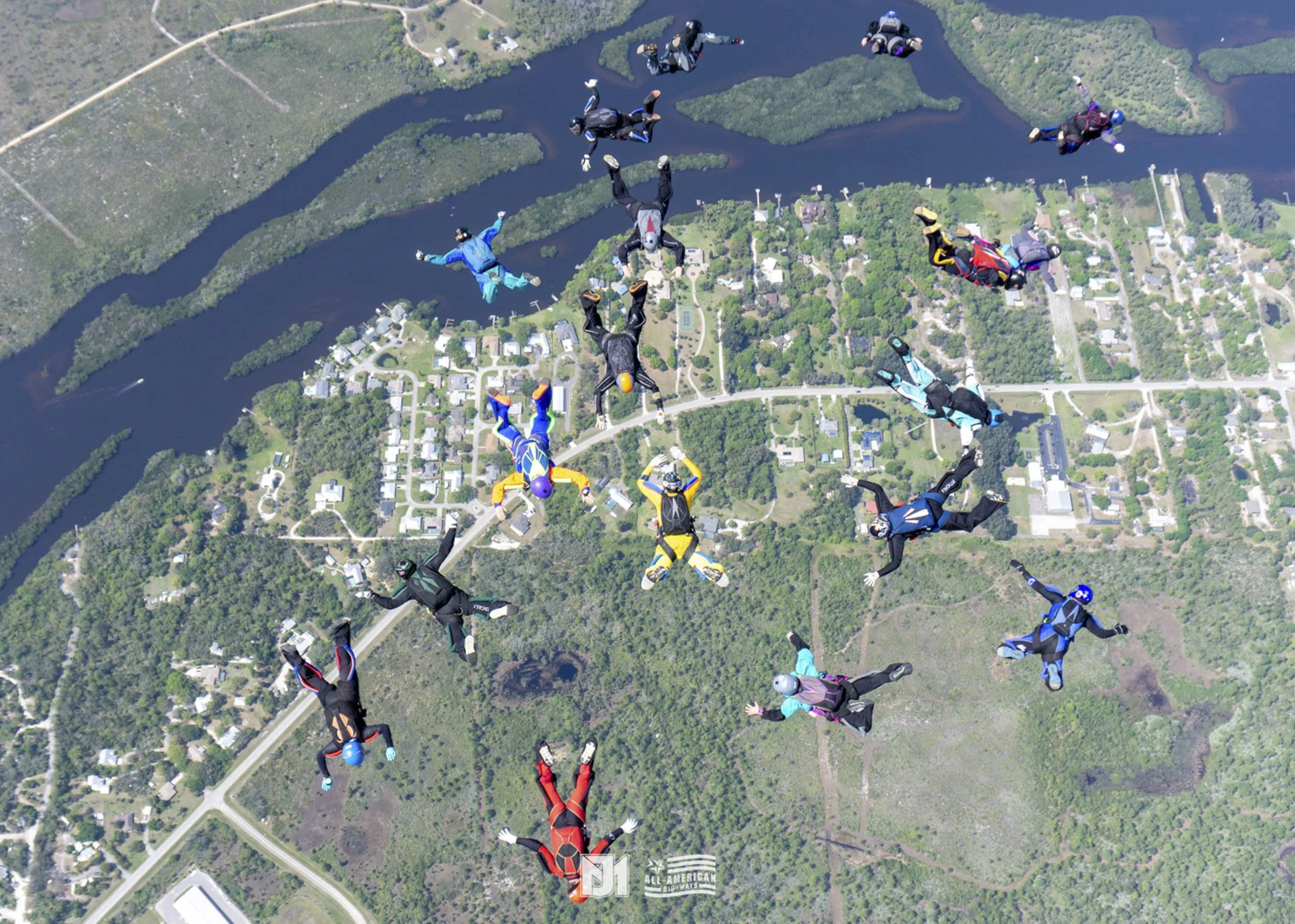 A group of skydivers free-falling over a landscape with lakes, trees, and buildings, wearing colorful jumpsuits and helmets.