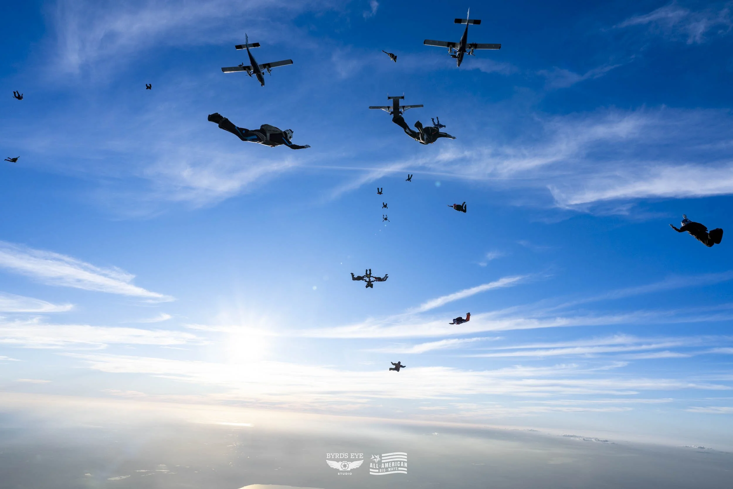 Multiple skydivers and airplanes mid-air against a blue sky with scattered clouds.