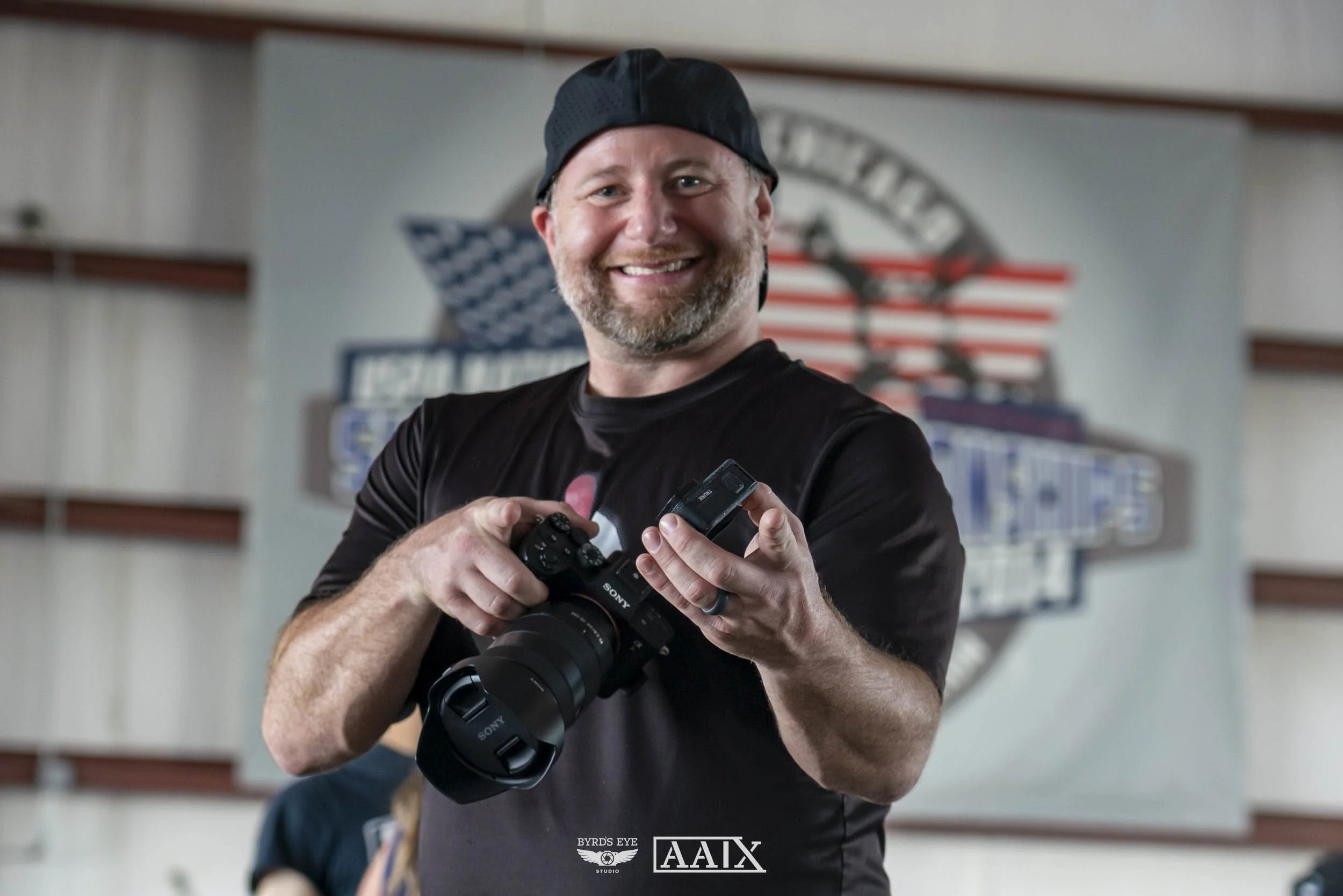 A smiling man standing indoors, holding a camera and a small device, with an American flag-themed logo in the background.