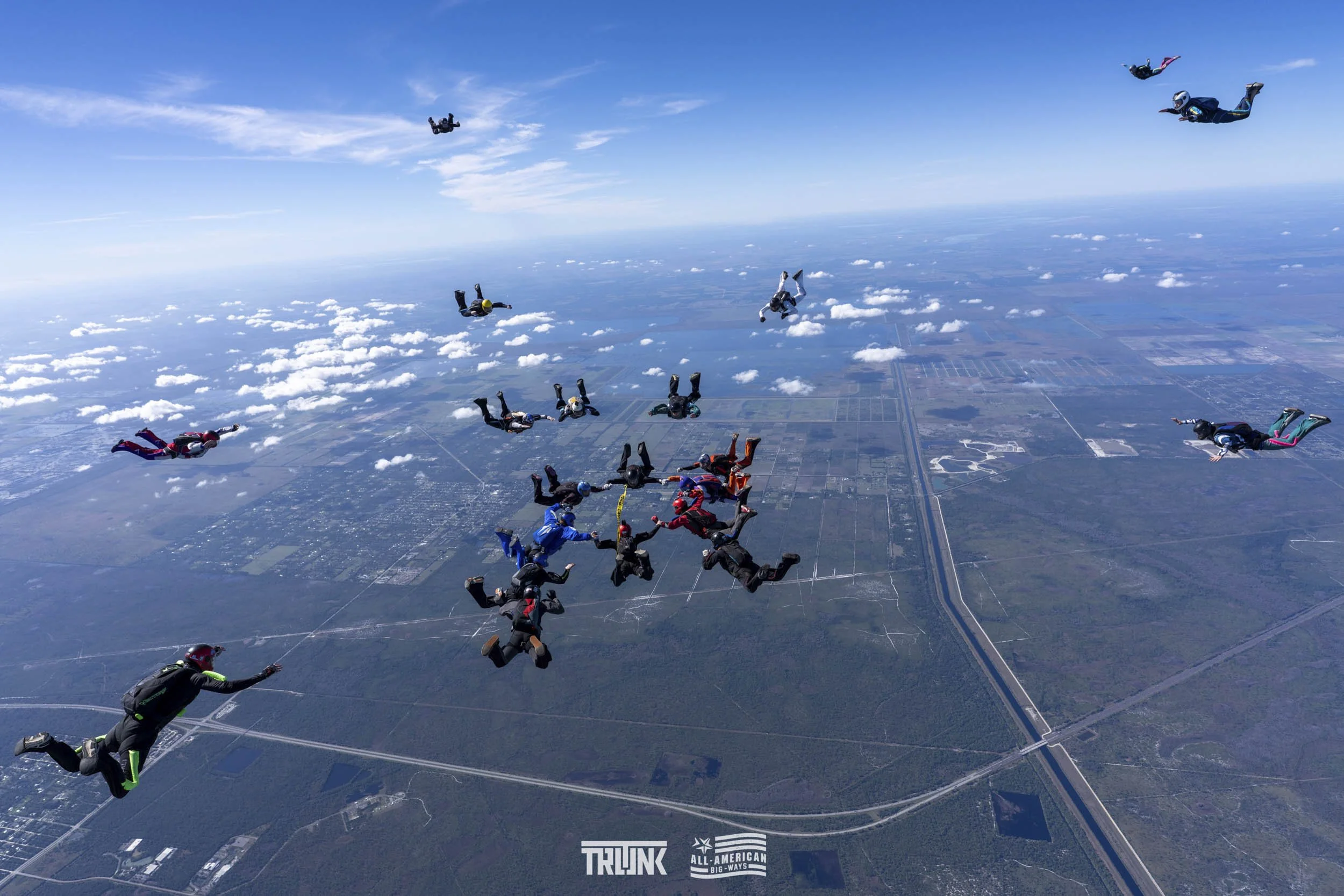 Group of skydivers in free fall over farmland and highways, with blue sky and clouds in the background.